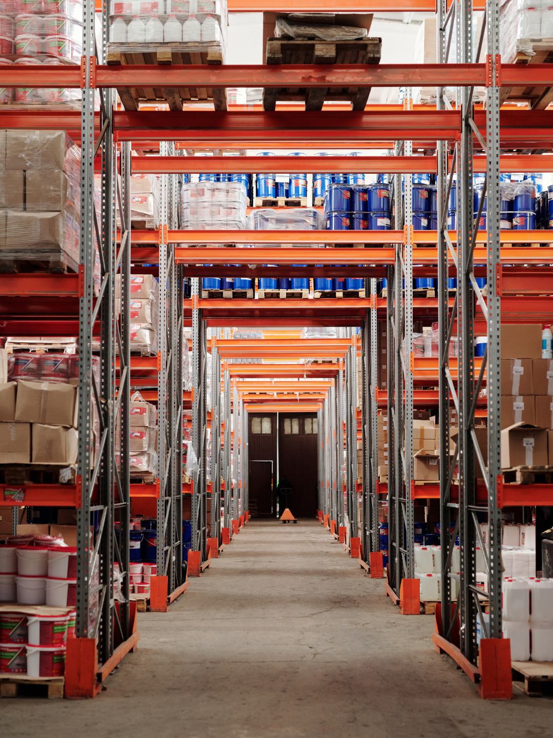 View of tall warehouse shelves stocked with paint cans, boxes, and supplies, with an open door at the end of the aisle.