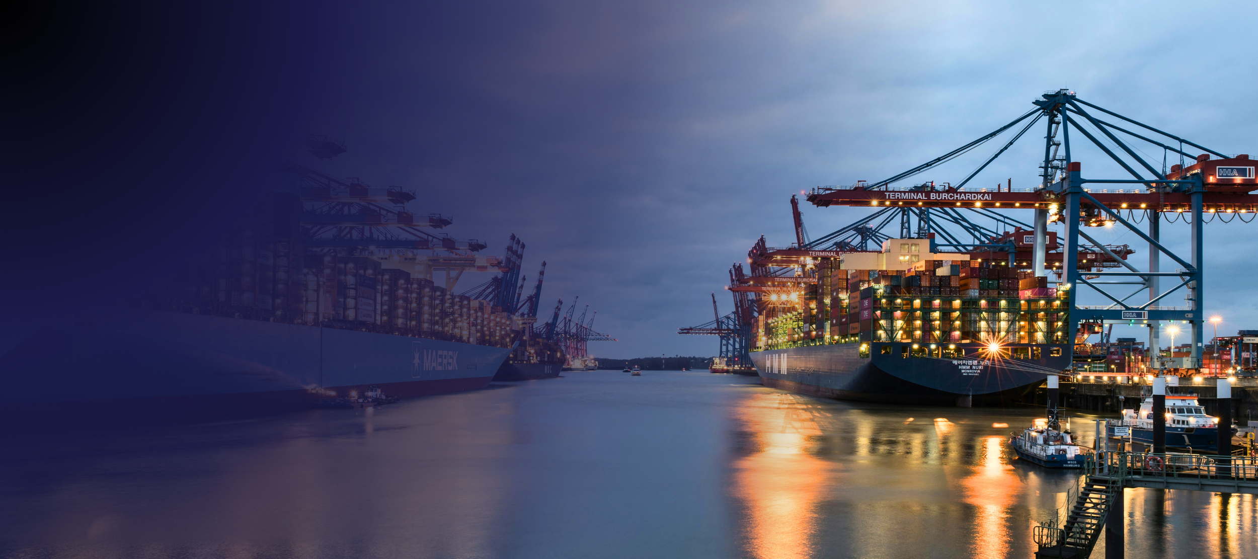 Cargo ships docked at a port with cranes loading containers during the evening with a partly cloudy sky.