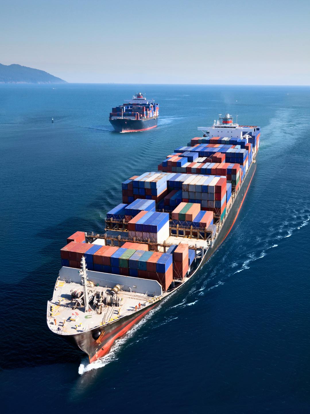 Large cargo ships transporting containers on the ocean, with a distant landmass visible in the background.