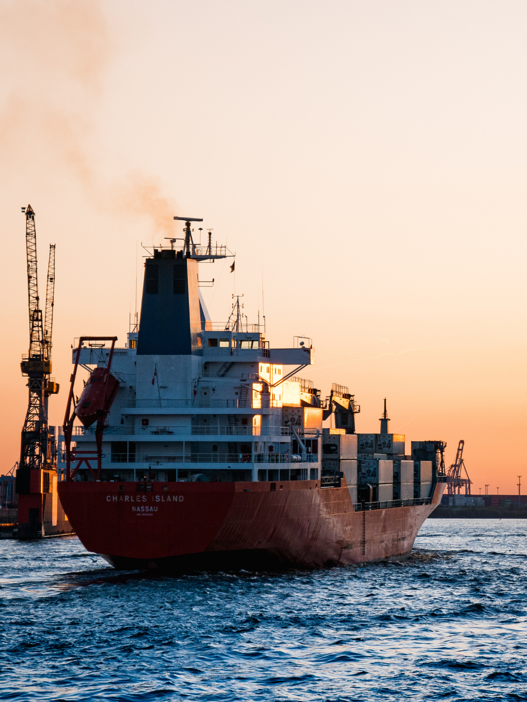 Cargo ship named Charles Island sailing in water during sunset with cranes and port in background.