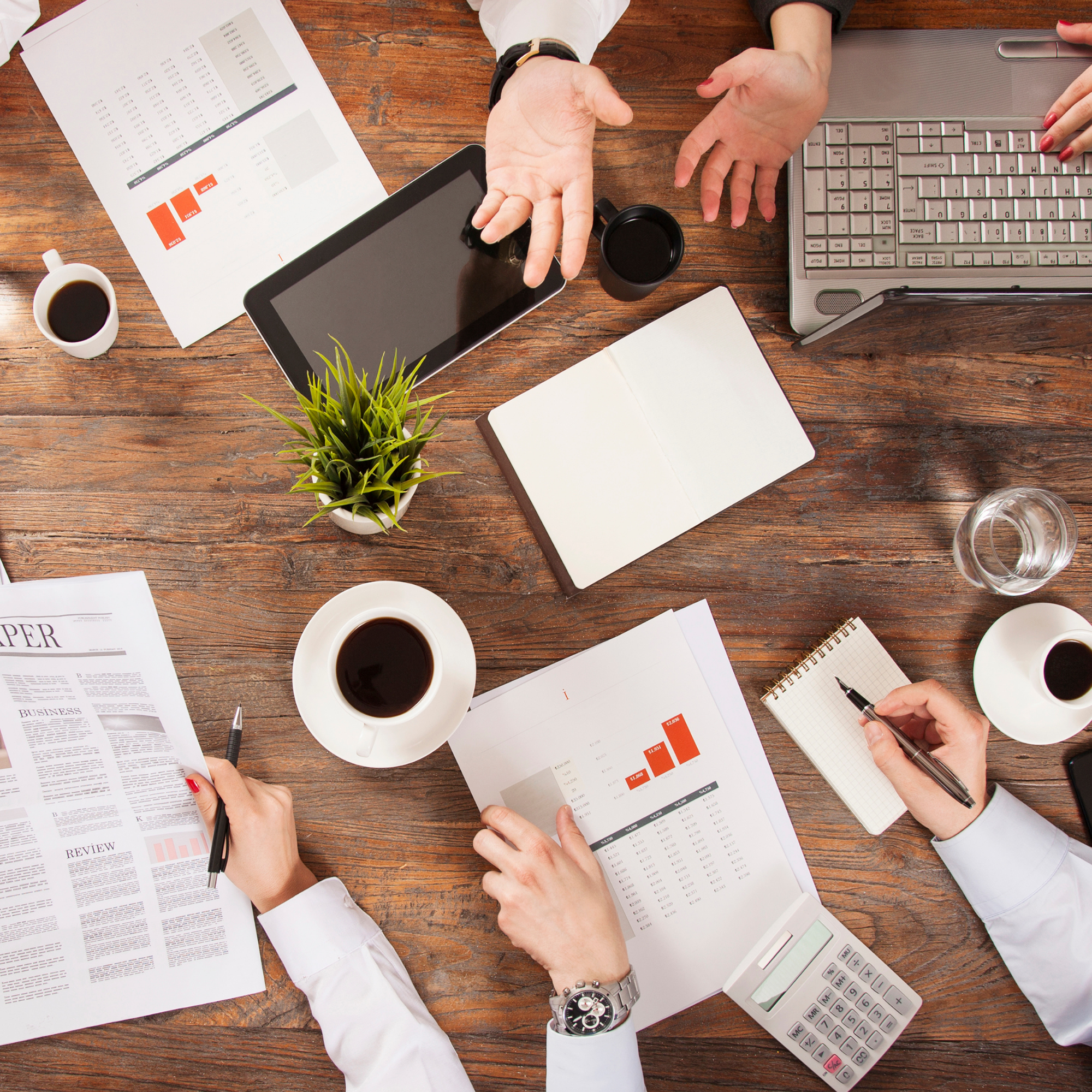 Multiple people working at a wooden desk with papers, notebooks, a calculator, cups of coffee, a potted plant, a smartphone, and a laptop, engaged in discussing financial or business data.
