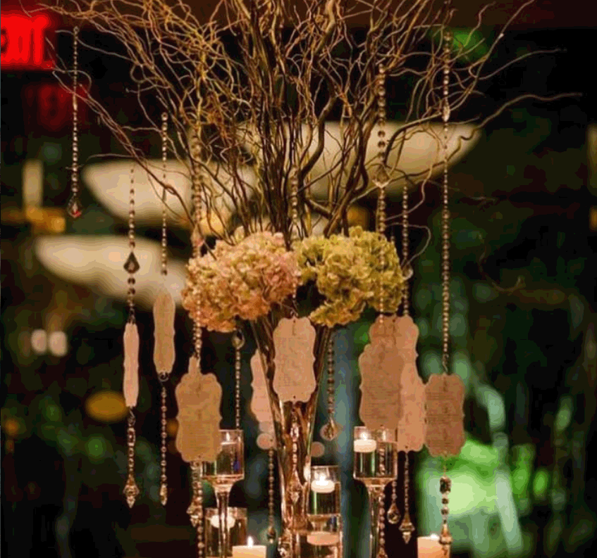 Decorative floral centerpiece with pink and green hydrangeas, surrounded by hanging tags and crystal bead strands, illuminated by small candles, in an elegant setting.