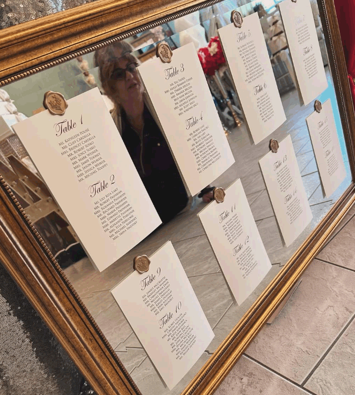 A framed mirror reflecting a woman and several printed seating cards for a wedding or event, each labeled with table numbers and guest names.