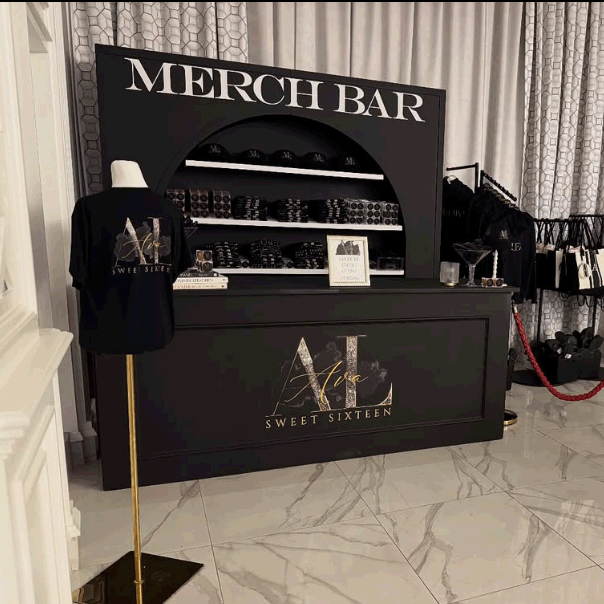 Merchandise bar with various items, a mannequin wearing a black T-shirt, and shelves with accessories, featuring branding for Aura Sweet Sixteen.