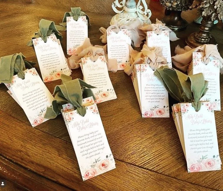 Set of personalized baby shower or wedding favor tags attached to cloth napkins, arranged on a wooden table, with decorative items in the background.