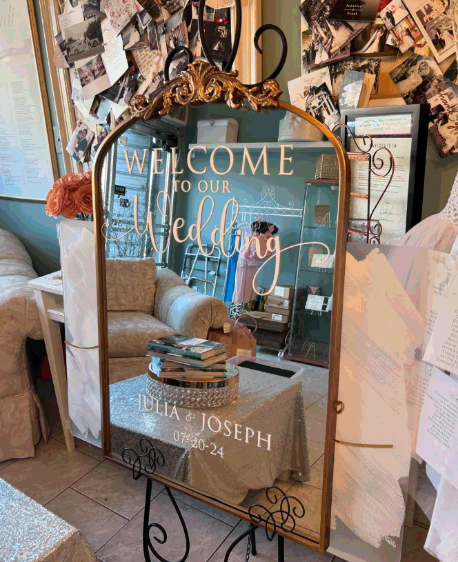 Decorative wedding welcome mirror sign with gold frame, reading 'Welcome to our Wedding, Julia & Joseph, 07-20-24', surrounded by wedding decor and photos.