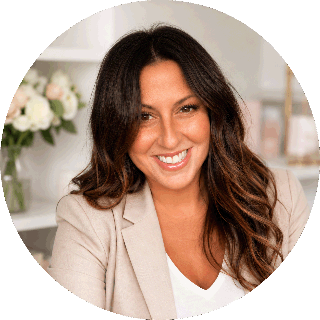 A woman with long, dark brown wavy hair smiling in an indoor setting with a blurred background of flowers and shelves.