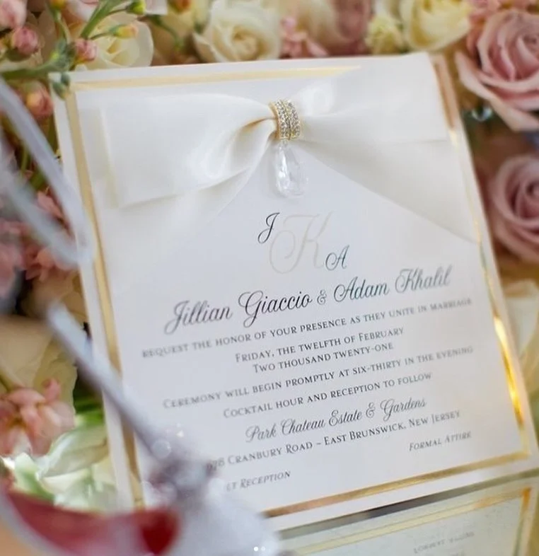 Wedding invitation with a white satin bow and diamond ring detail, surrounded by pink and white roses.