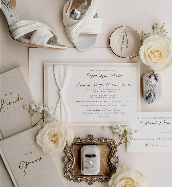 Wedding invitation surrounded by white roses, a pair of white heels, a gold-rimmed jewelry dish, and a vintage ring box with rings, set on a white background.