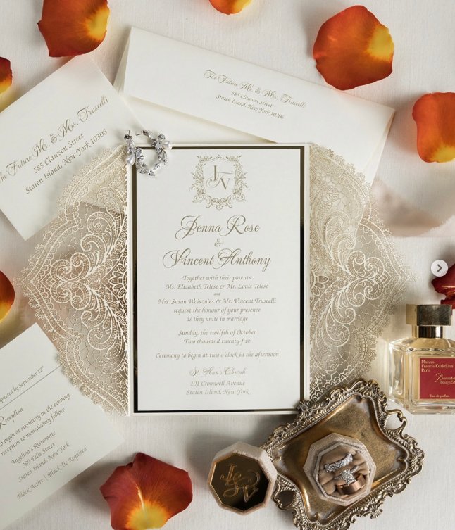 Wedding invitation with rings, jewelry, and perfume on a lace doily, surrounded by rose petals.