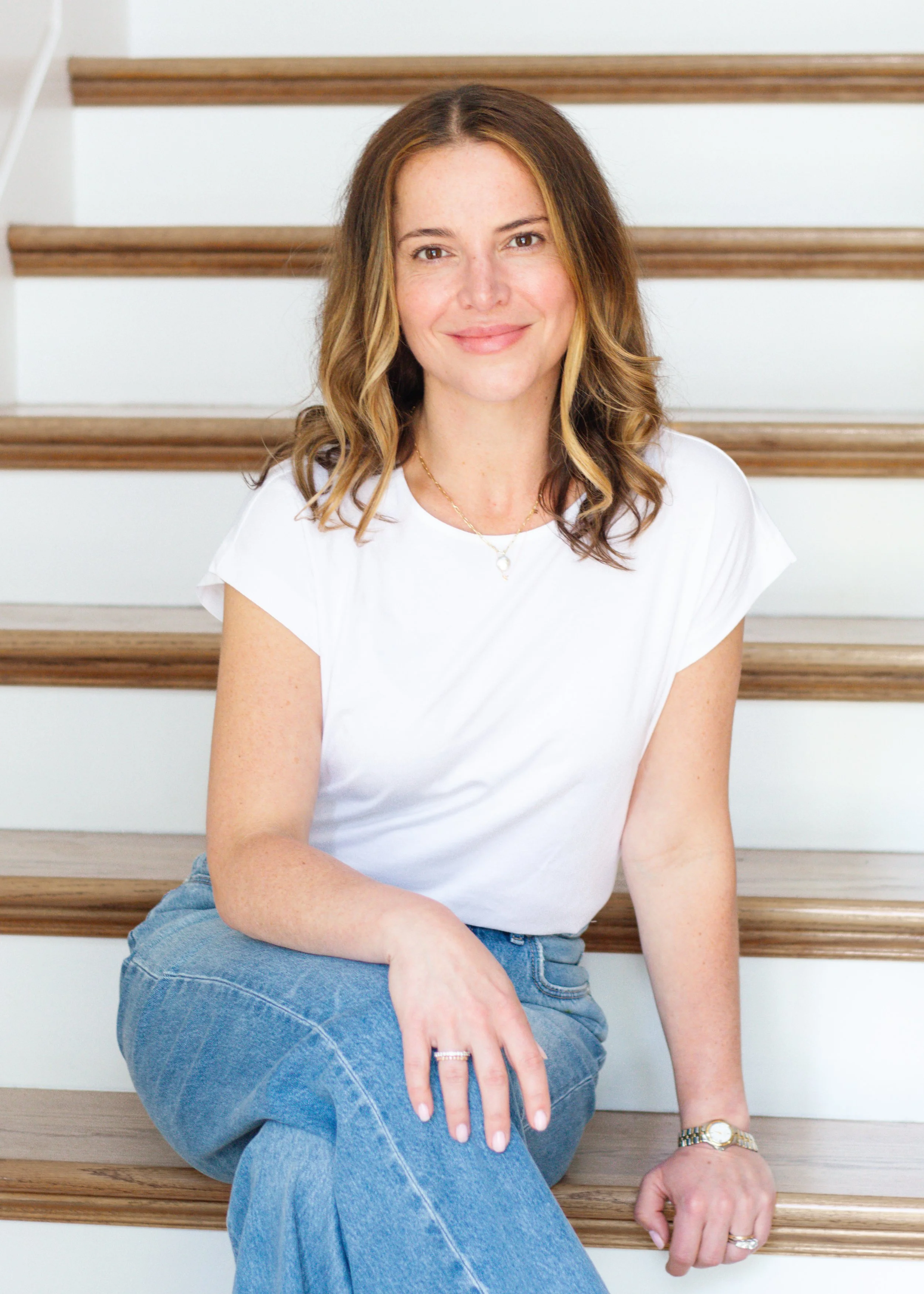 A woman with shoulder-length, wavy, light brown hair sitting on wooden stairs, wearing a white t-shirt and blue jeans, smiling at the camera.