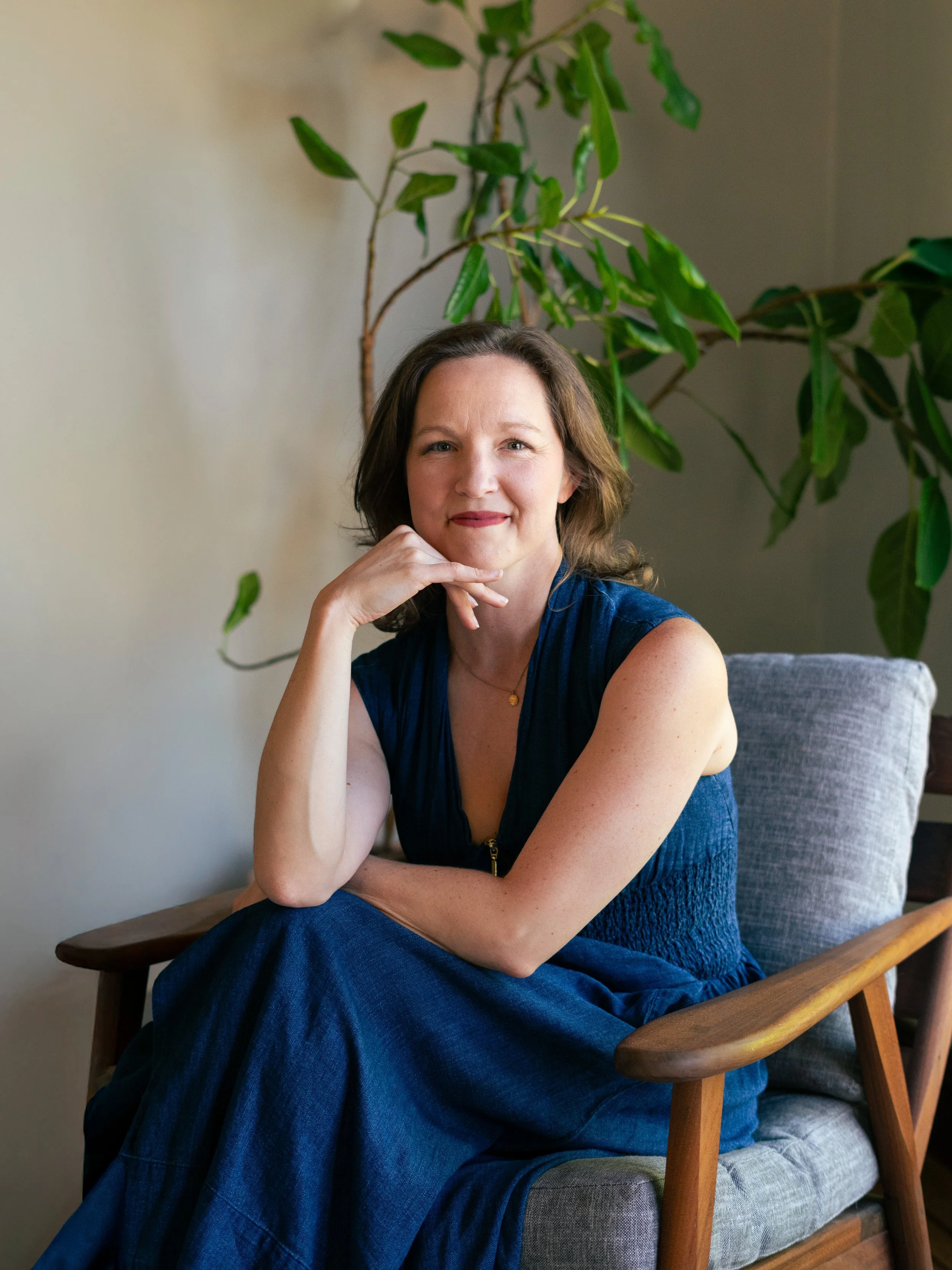 A woman sitting on a grey upholstered chair with wooden armrests, resting her chin on her hand, smiling at the camera. She has shoulder-length brown hair, is wearing a sleeveless dark blue dress, and a gold necklace. Behind her, there is a large green plant with broad leaves, and the background features a beige wall.