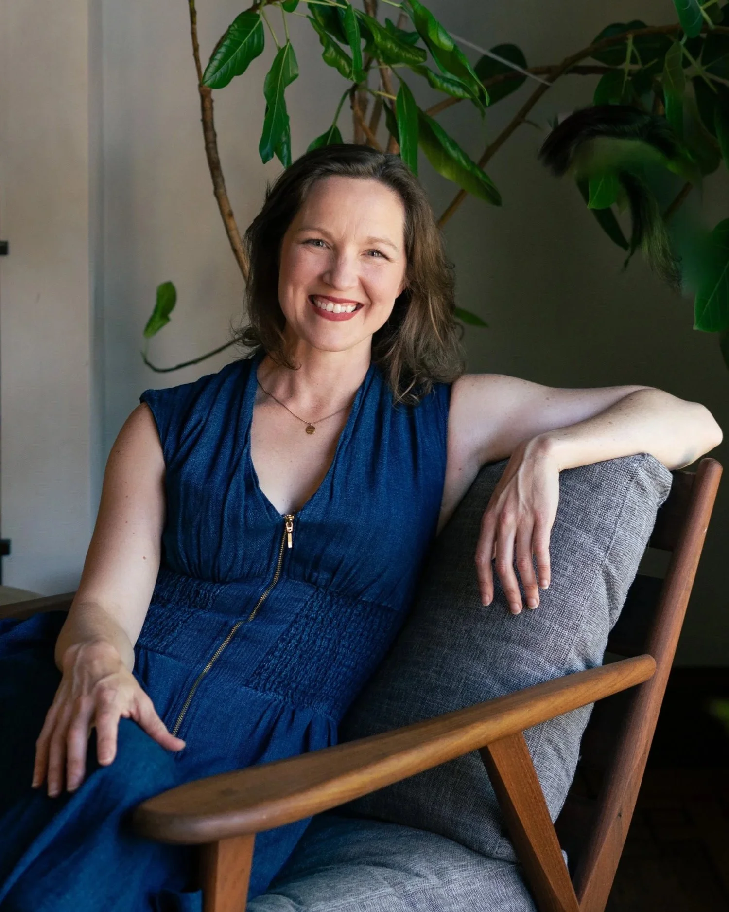A woman with shoulder-length brown hair, wearing a blue sleeveless top and a gold necklace, sitting on a chair with a wooden frame and armrest, smiling at the camera in front of a large leafy green plant.