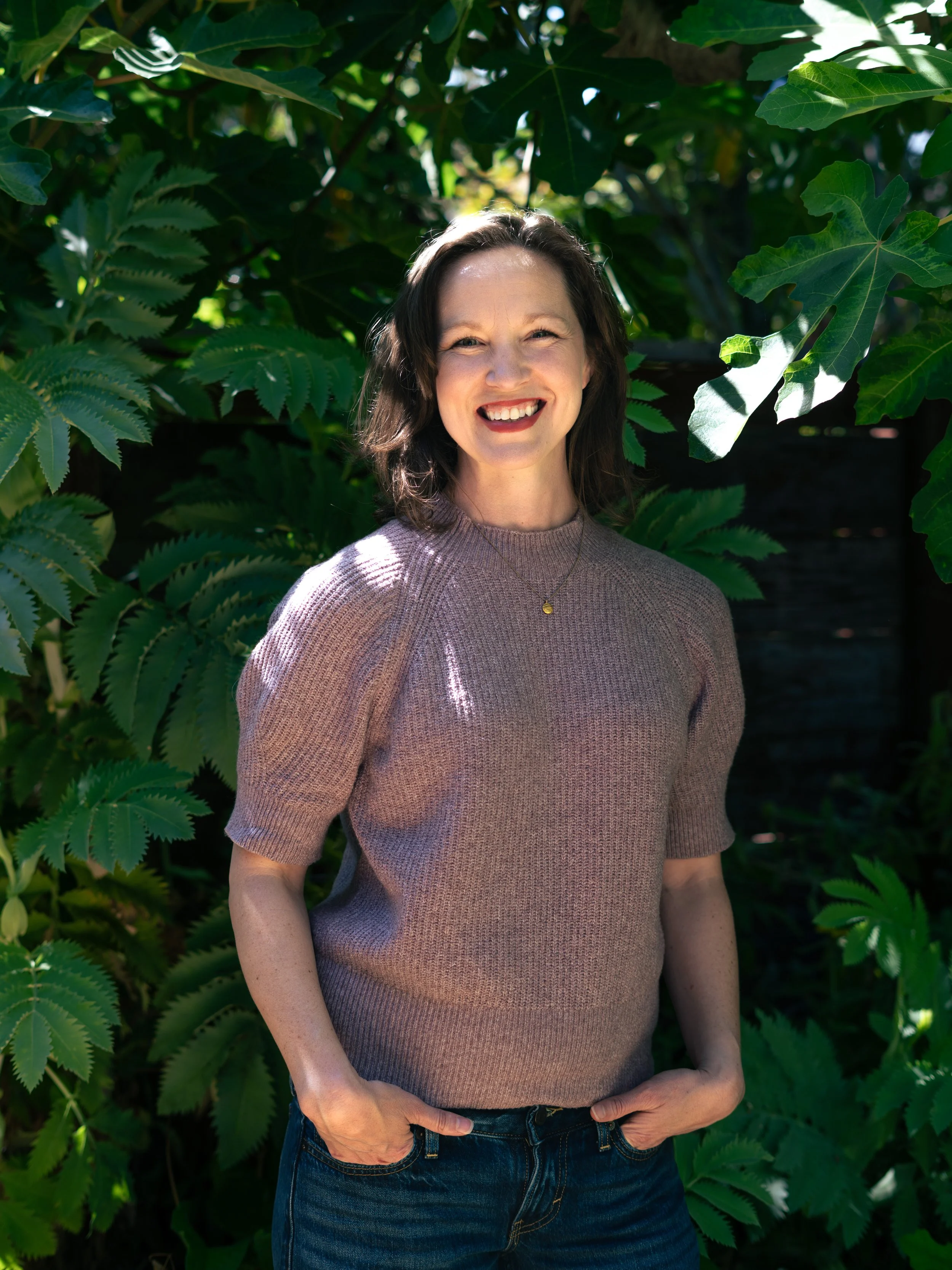 Smiling woman standing outdoors between green leafy trees, wearing a beige sweater and blue jeans.