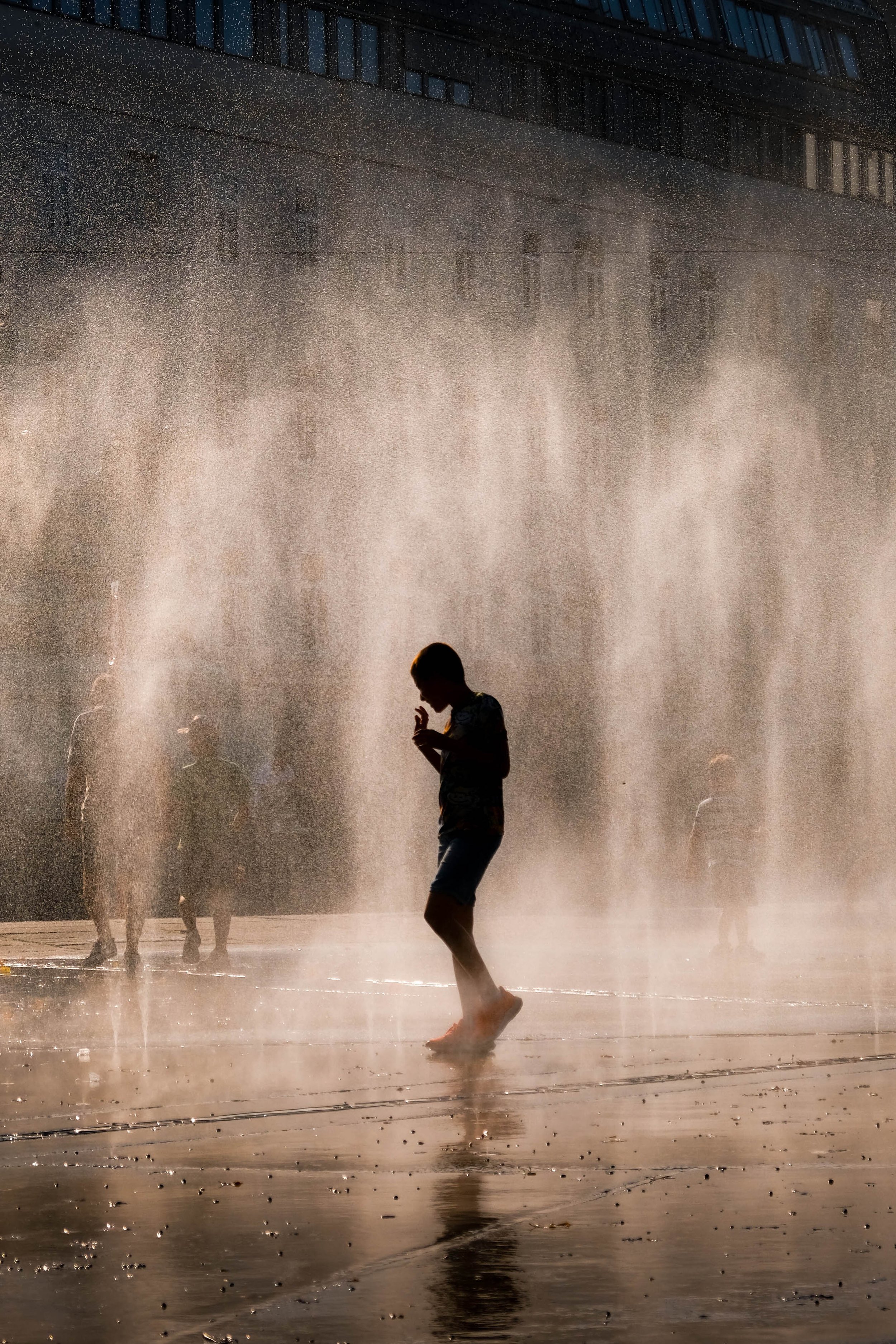 Une silhouette de personne qui marche au milieu d'une fontaine d'eau en spray, avec d'autres personnes en arrière-plan, dans une ville la nuit.