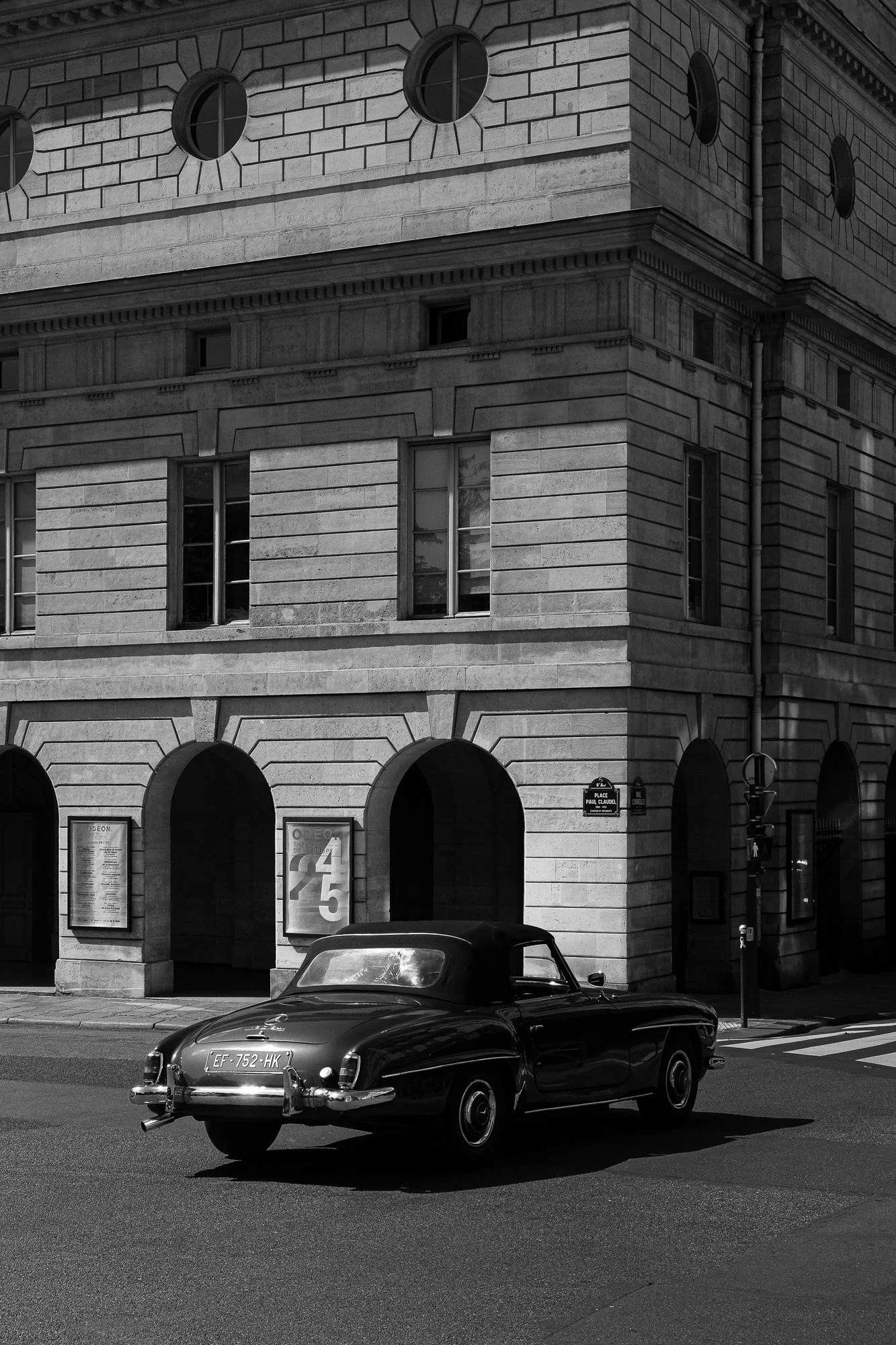 Une rue avec une voiture classique noire garée devant un bâtiment en pierre avec des fenêtres rectangulaires et des arches. La façade du bâtiment comporte plusieurs fenêtres et un panneau d'affichage, avec une enseigne indiquant la place Paul Claudel