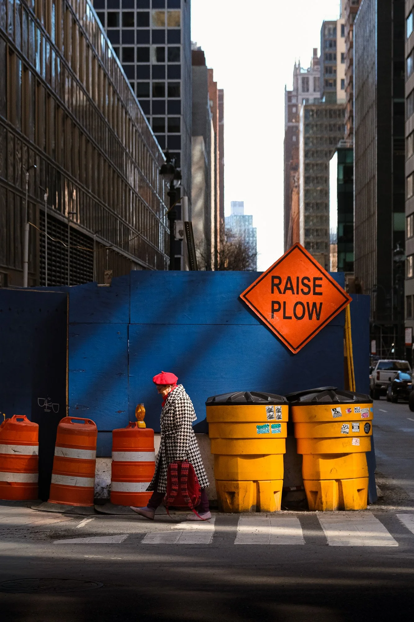 Femme marchant à travers un chantier de construction en ville, portant un manteau à motifs et une casquette rouge. Signaleur orange 'Raise Plow' et barrières orange et jaunes. Gratte-ciel en arrière-plan.