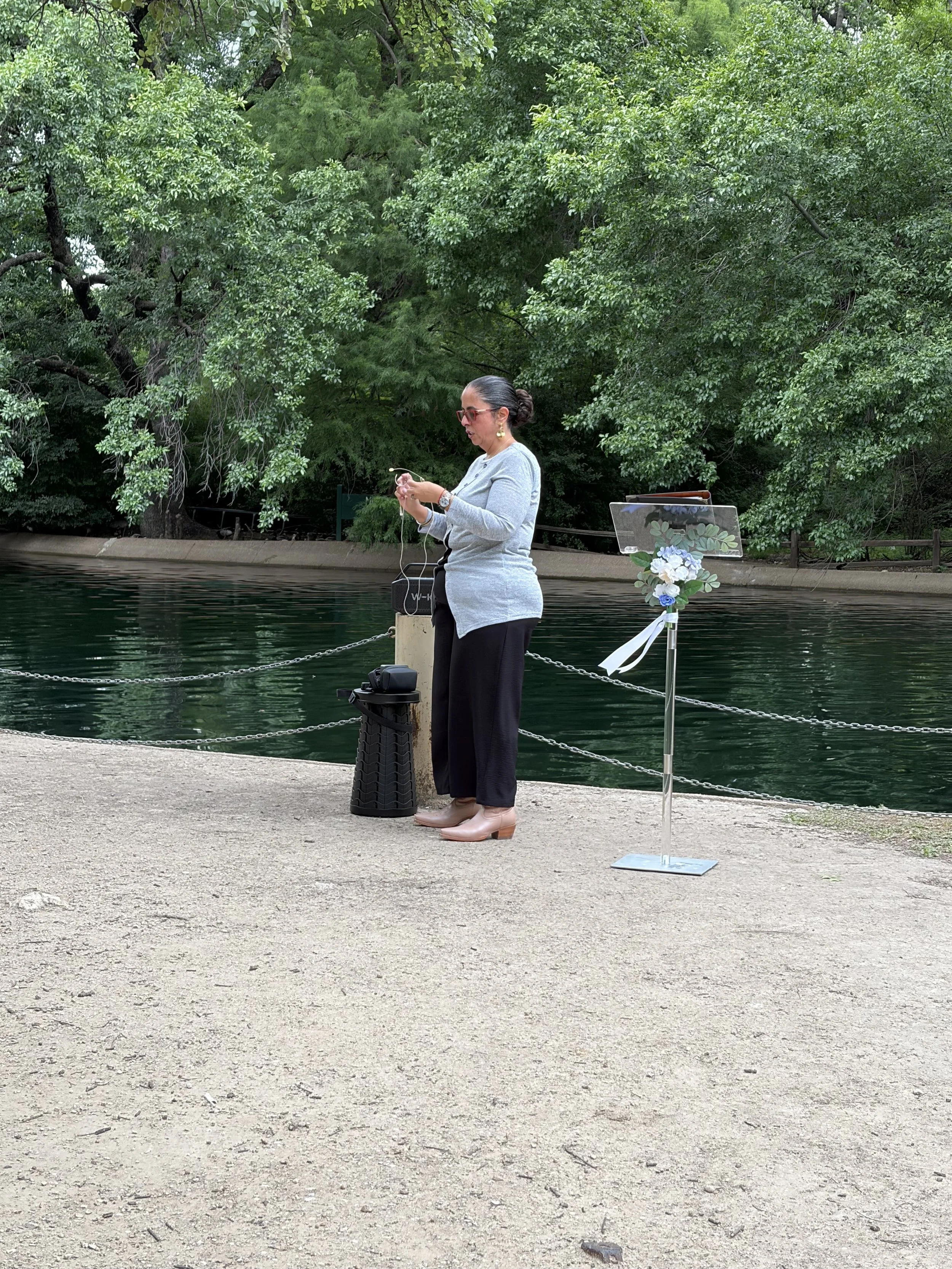 A woman in gray sweater, black pants, and beige boots standing by a body of water, tying her shoelace. There is a small table with flowers and a ribbon in front of her, and a stand with a clear plastic cover and items inside, near her.