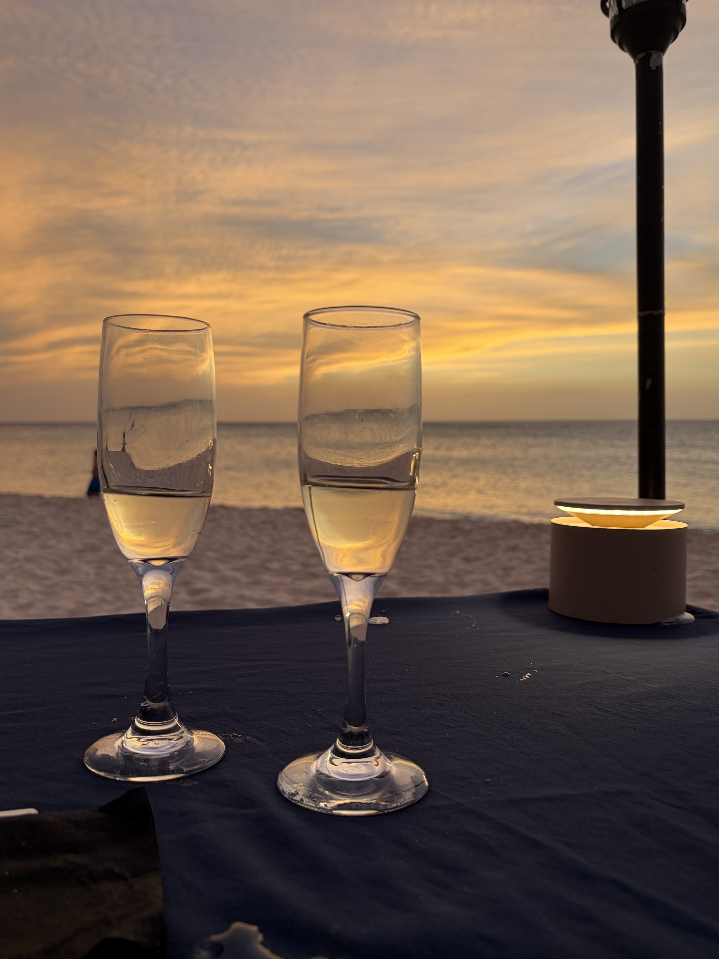 Two champagne glasses filled with a golden beverage on a black table, with a sunset sky and ocean in the background, and a table umbrella on the right.