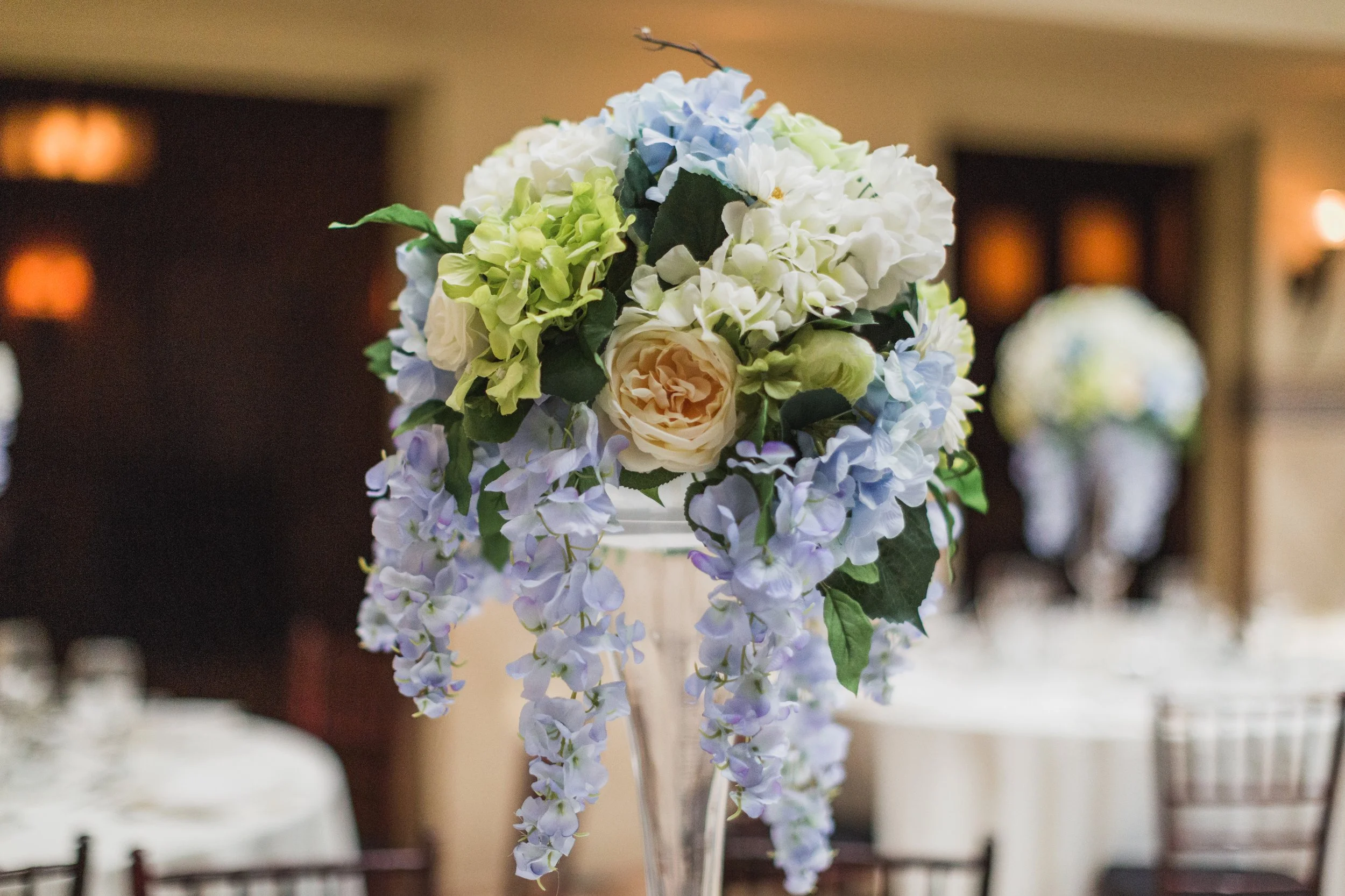 A floral arrangement of white, cream, and light blue hydrangeas and roses, displayed in a glass vase on a table indoors.