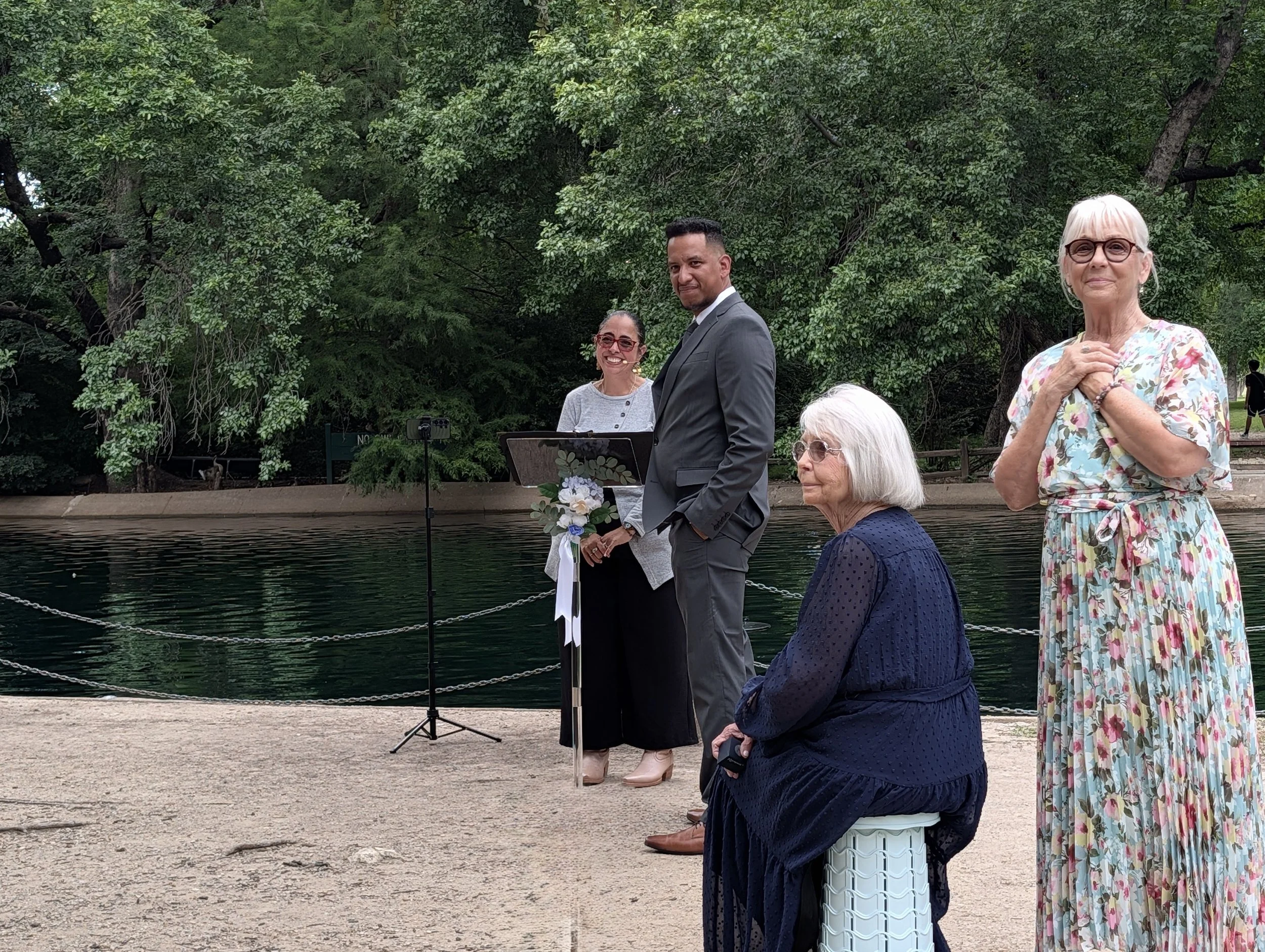 People gathered outdoors near a water feature with a man in a gray suit, a woman with white hair and glasses, and others in casual attire, surrounded by green trees.