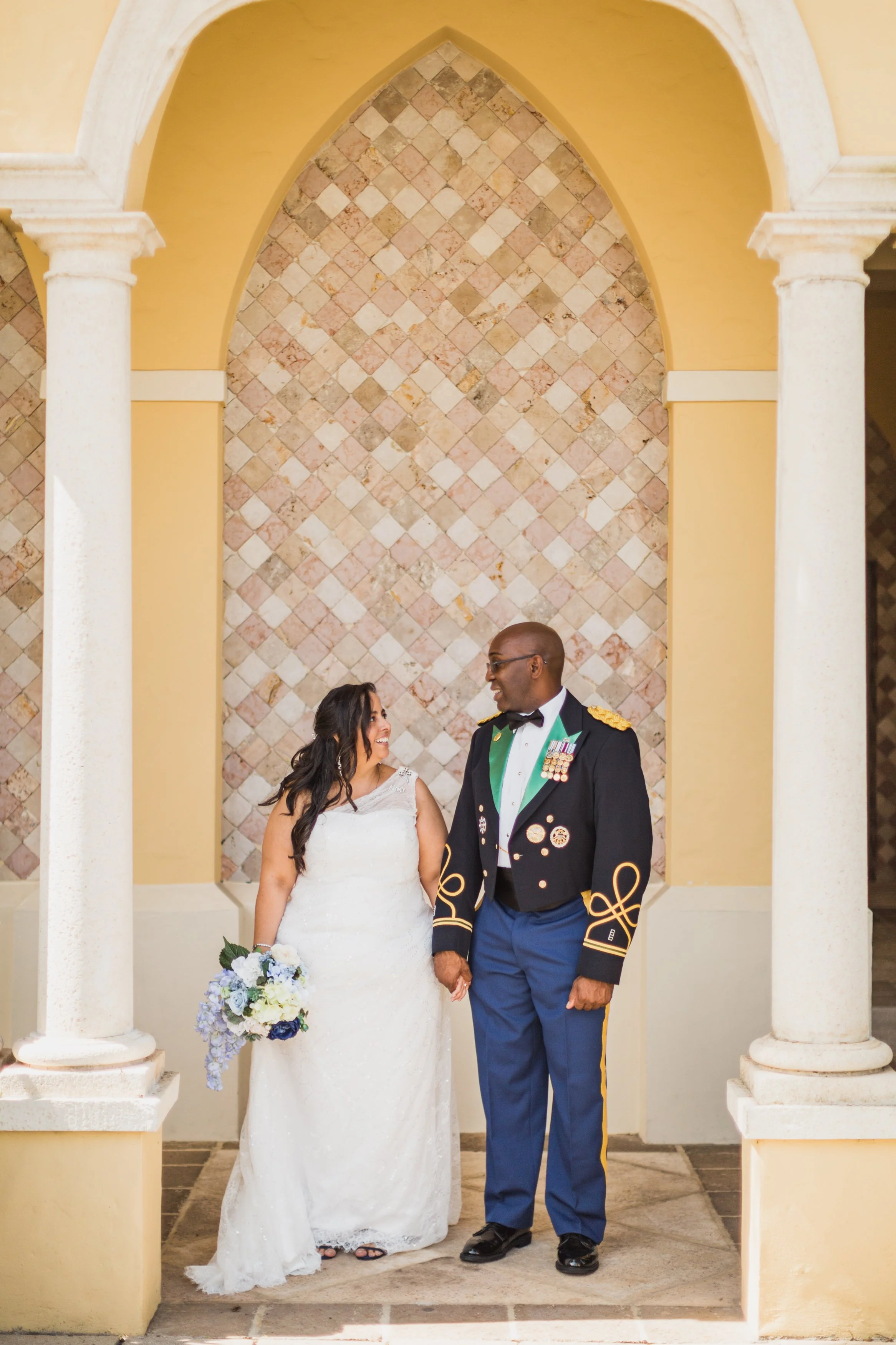 A bride and a groom stand holding hands in front of a decorative stone wall, underneath a yellow archway with white columns.