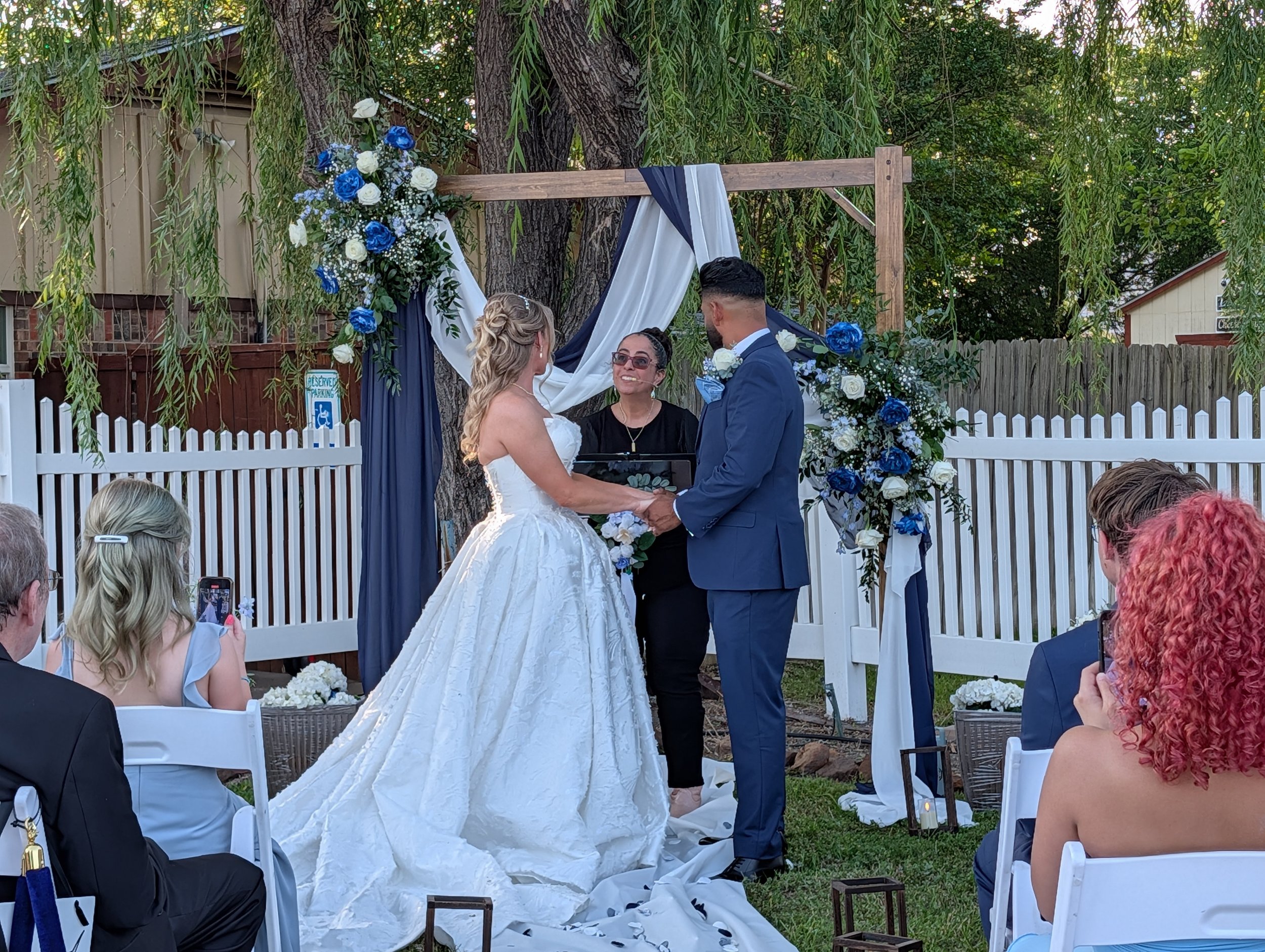 A wedding ceremony with a bride and groom holding hands, standing under a decorated arch with greenery and blue and white flowers, outside in a garden setting, with guests seated in front.