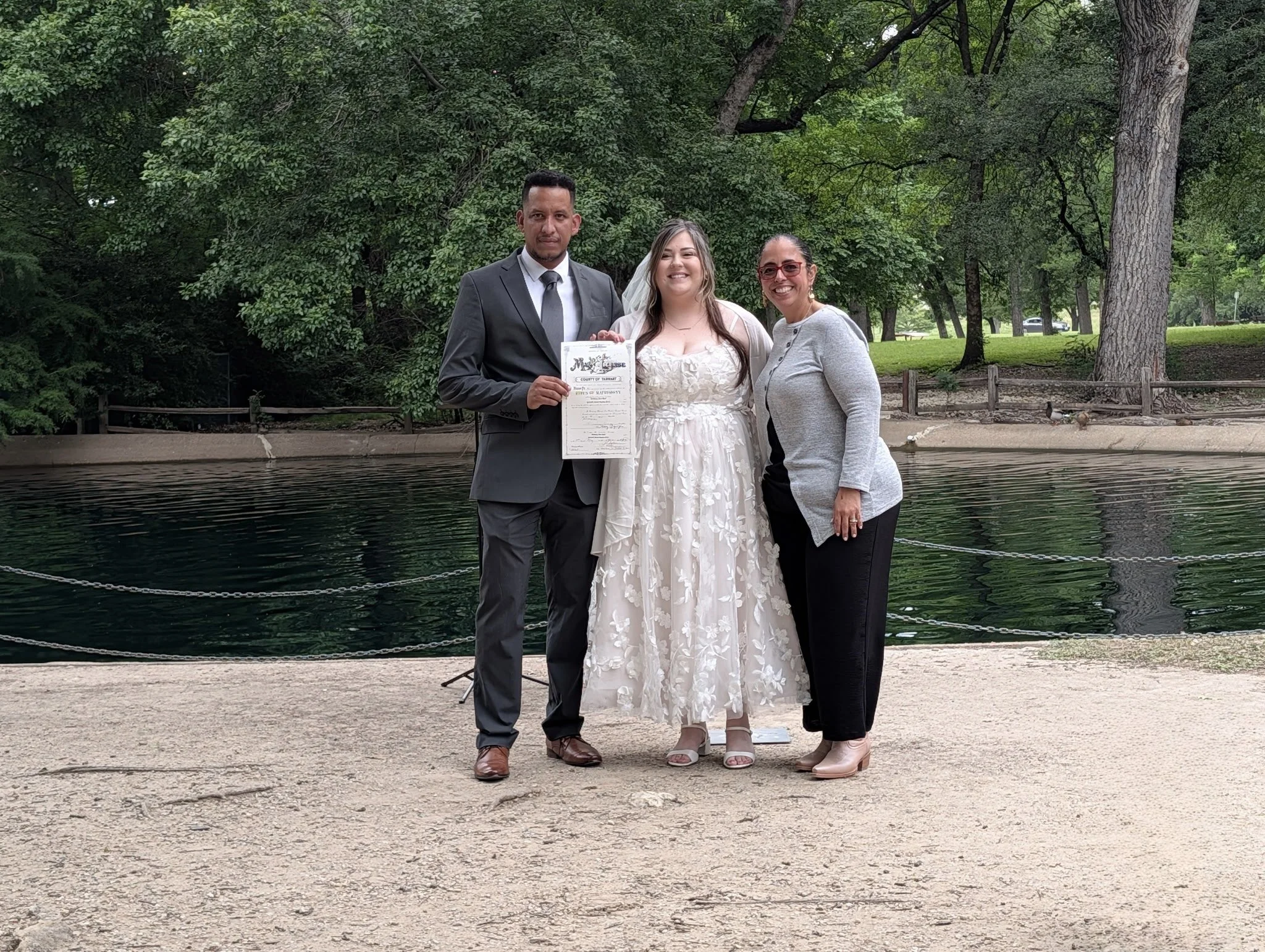 Three people posing outdoors by the water in a park, with tall trees in the background. The person on the left is wearing a suit and holding a certificate, the person in the middle is in a white dress, and the person on the right is in a gray top and black pants.