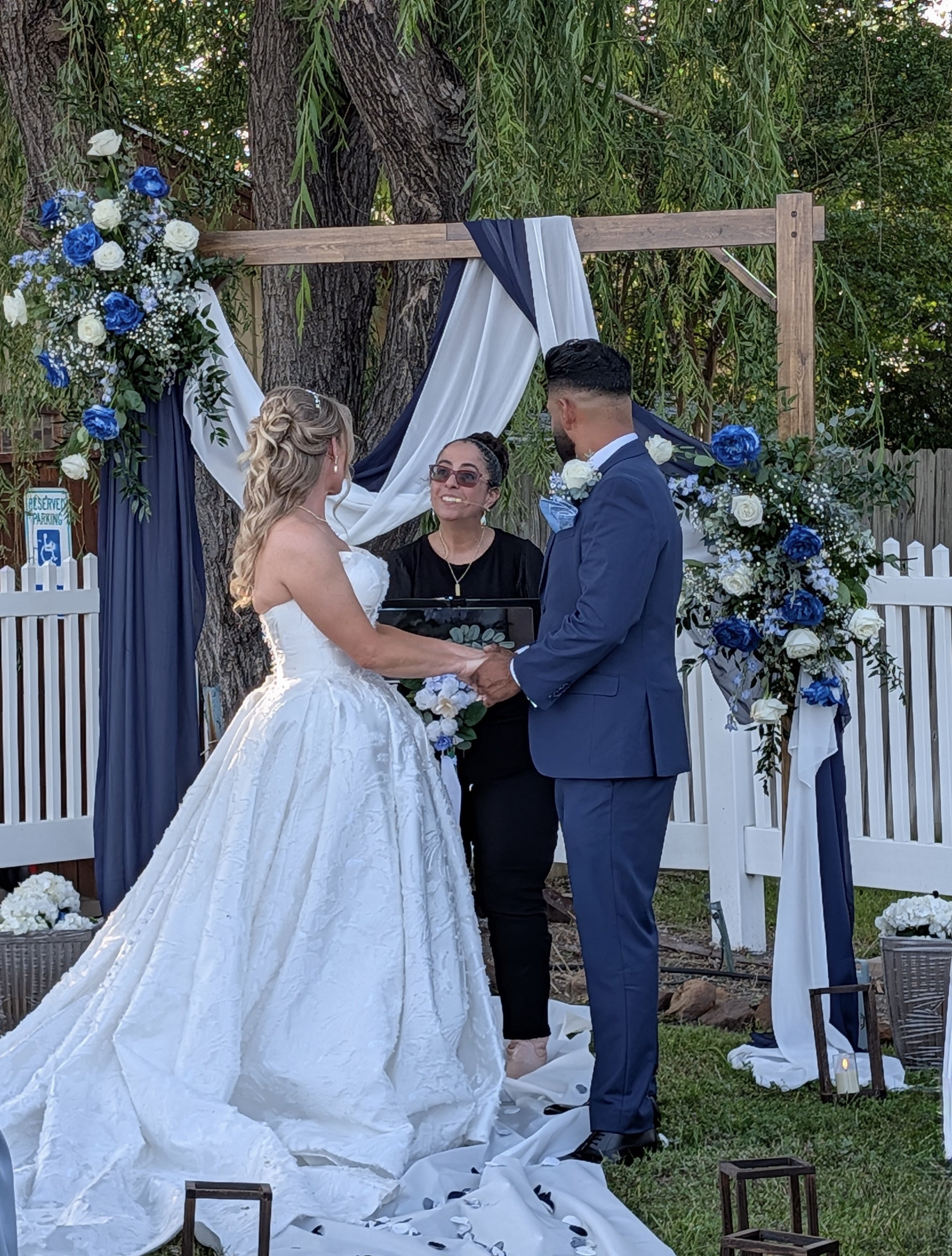 A wedding ceremony outdoors with a bride and groom holding hands and facing each other, an officiant standing in front of them, and a decorated arch with blue and white flowers behind them.