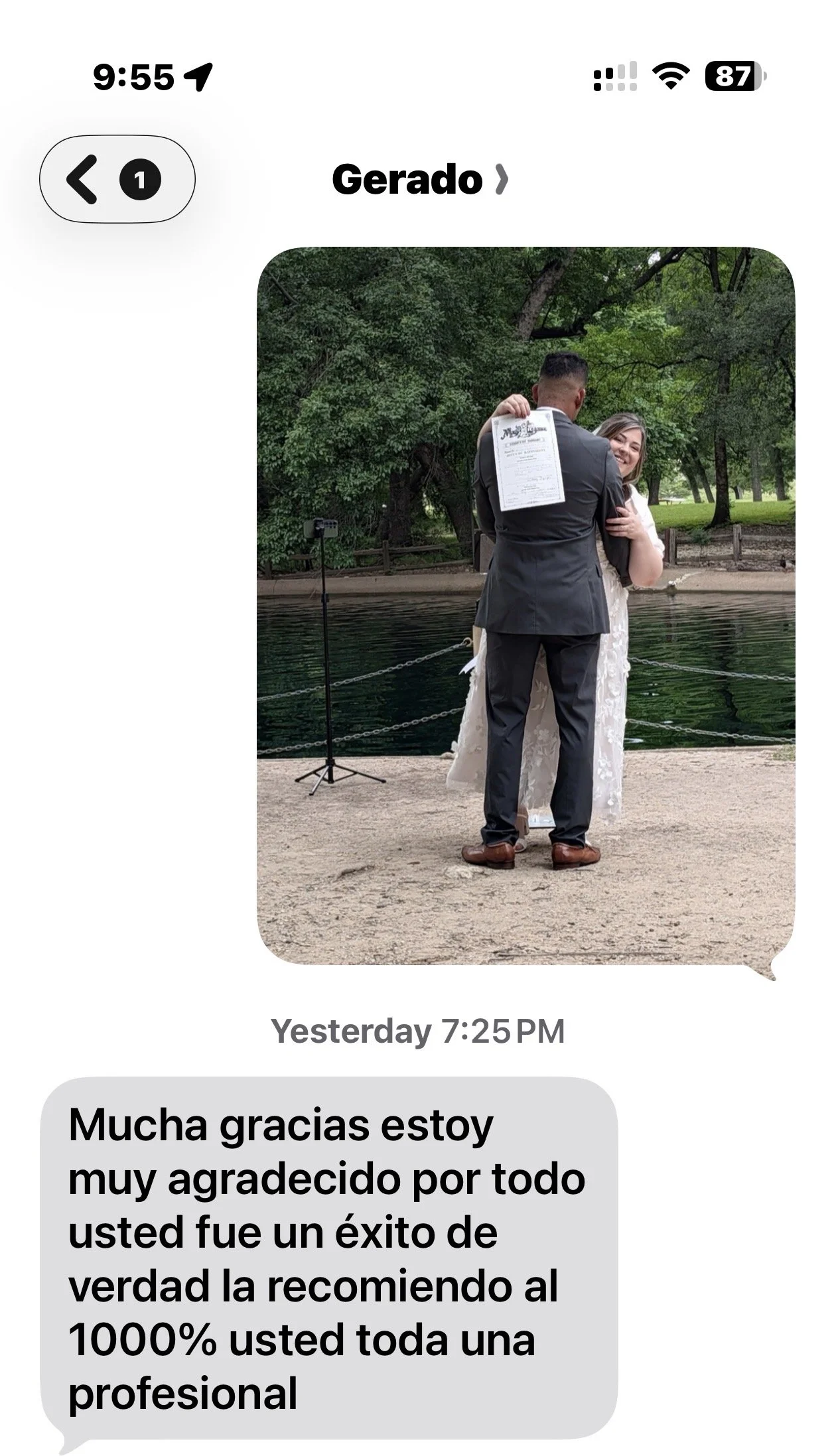 A couple is embracing outdoors near a body of water, with the woman smiling and the man holding a paper behind his back in a park-like setting with trees in the background.
