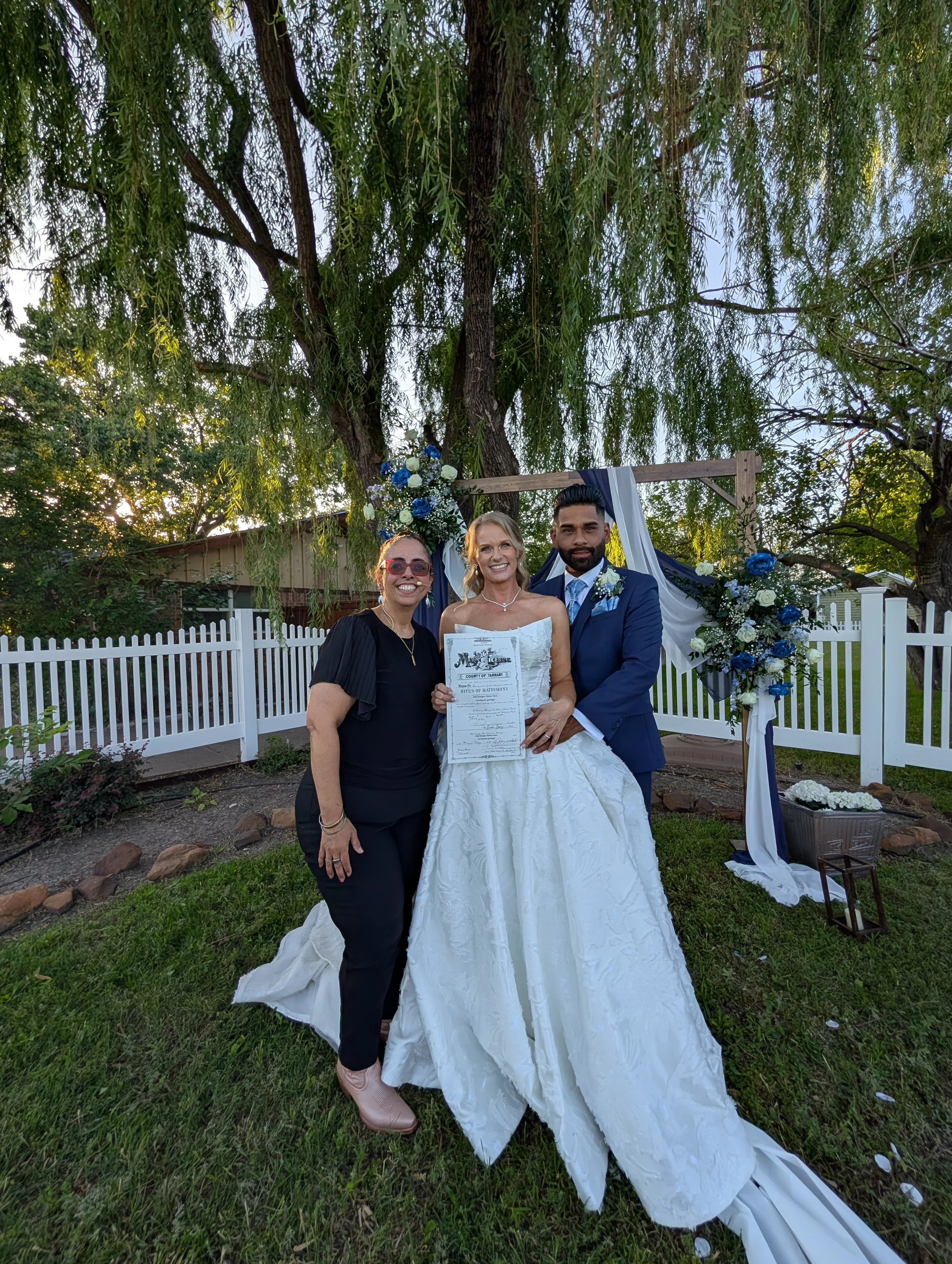 Wedding couple, bride and groom in front of wooden arch with blue and white flowing fabric and flowers. Wedding officiant is posing with the beautiful bride and groom.
