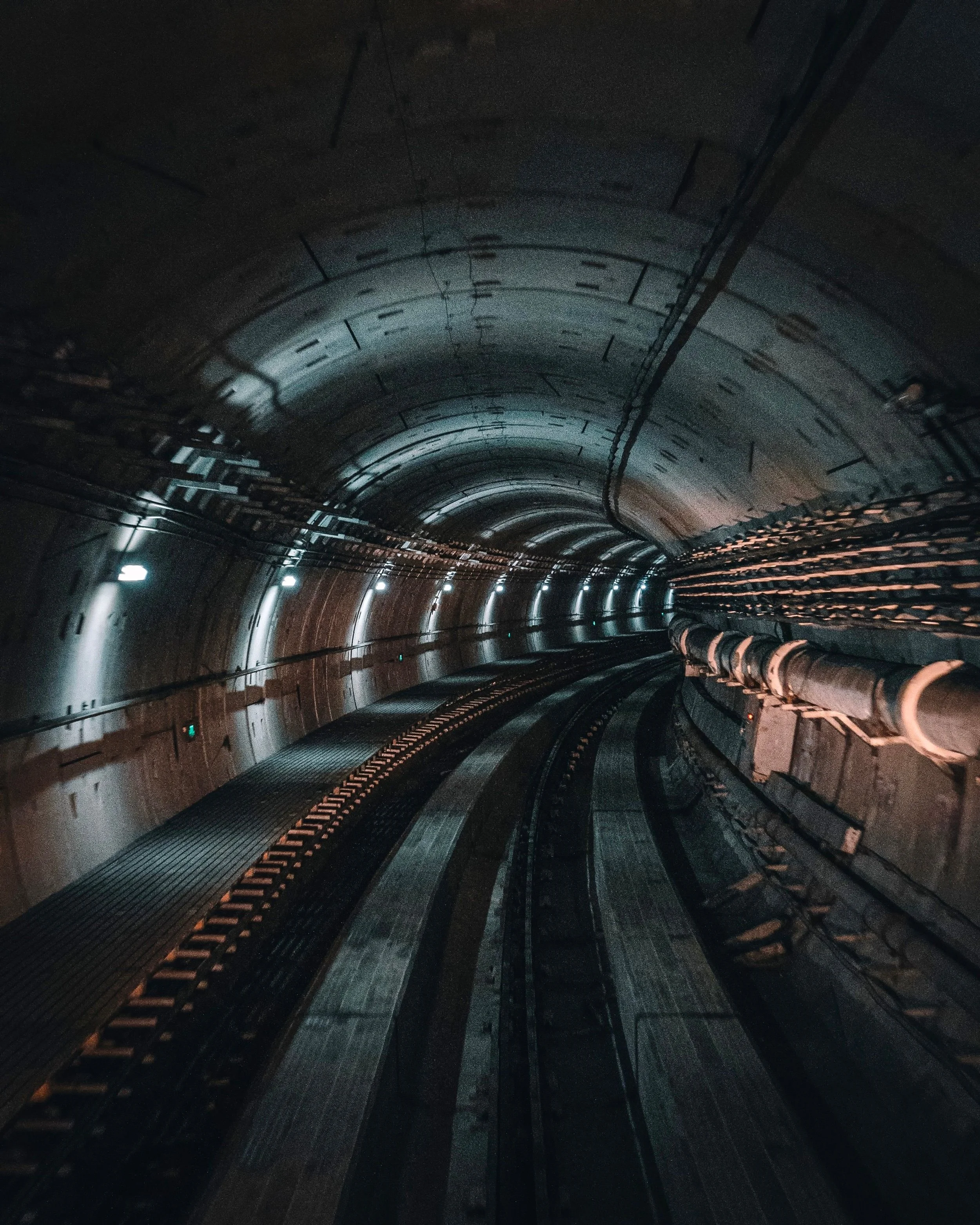 Dark tunnel with lit lights along the sides and visible piping and wiring on the ceiling.