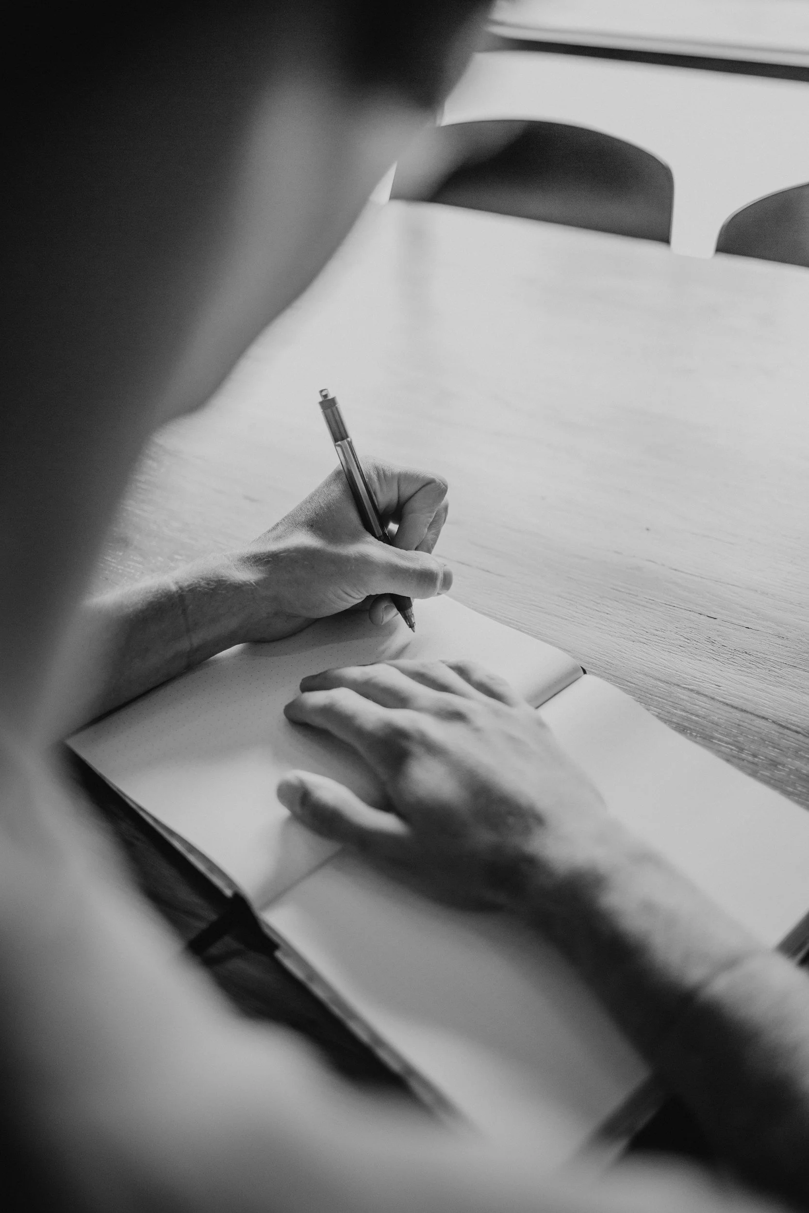 A person is writing in a notebook at a wooden table, with a hand resting on the paper and a pen in hand.