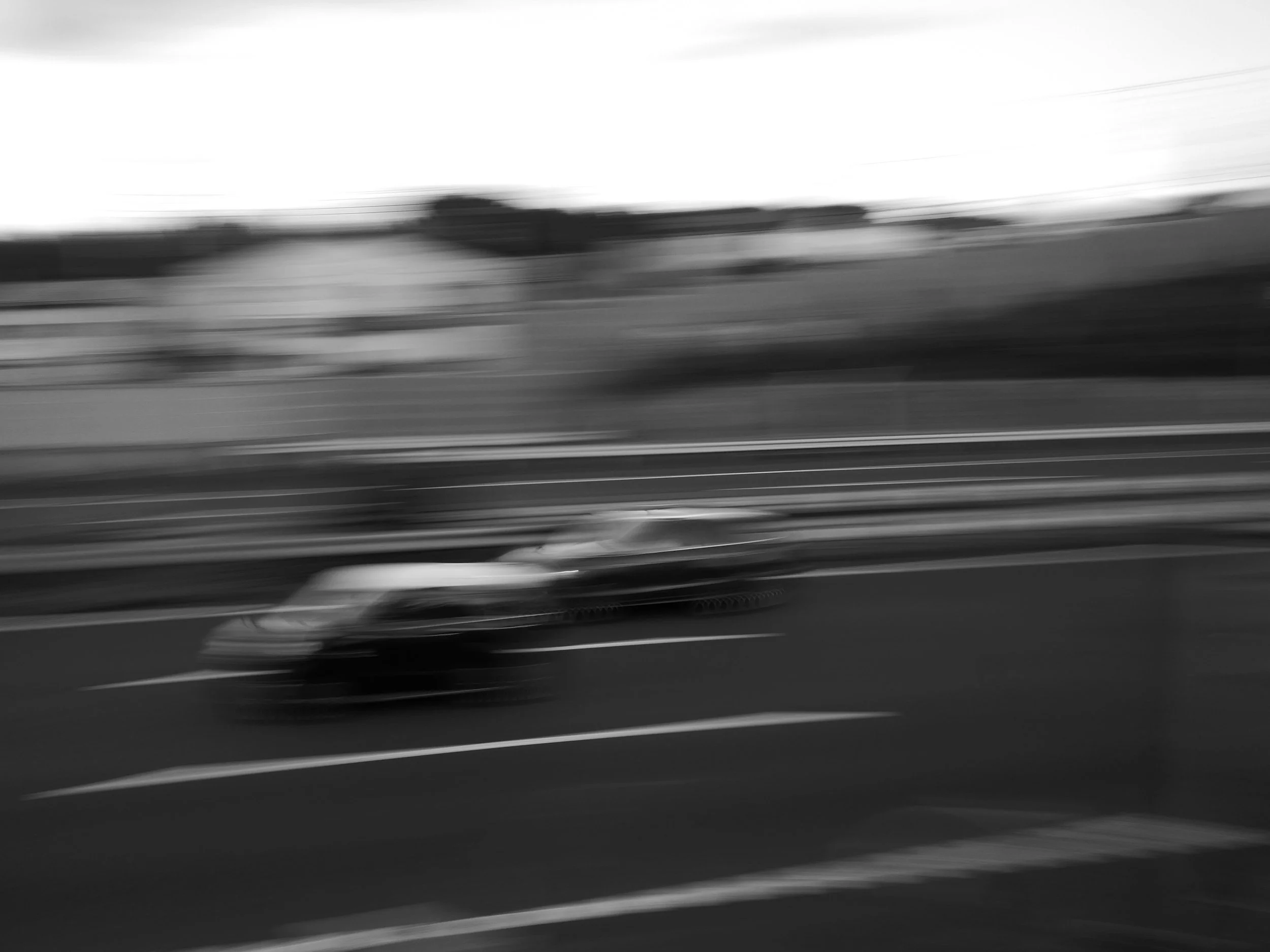 Blurred black and white photo of a moving vehicle on a road, with visible lane markings and a landscape with trees and power lines in the background.
