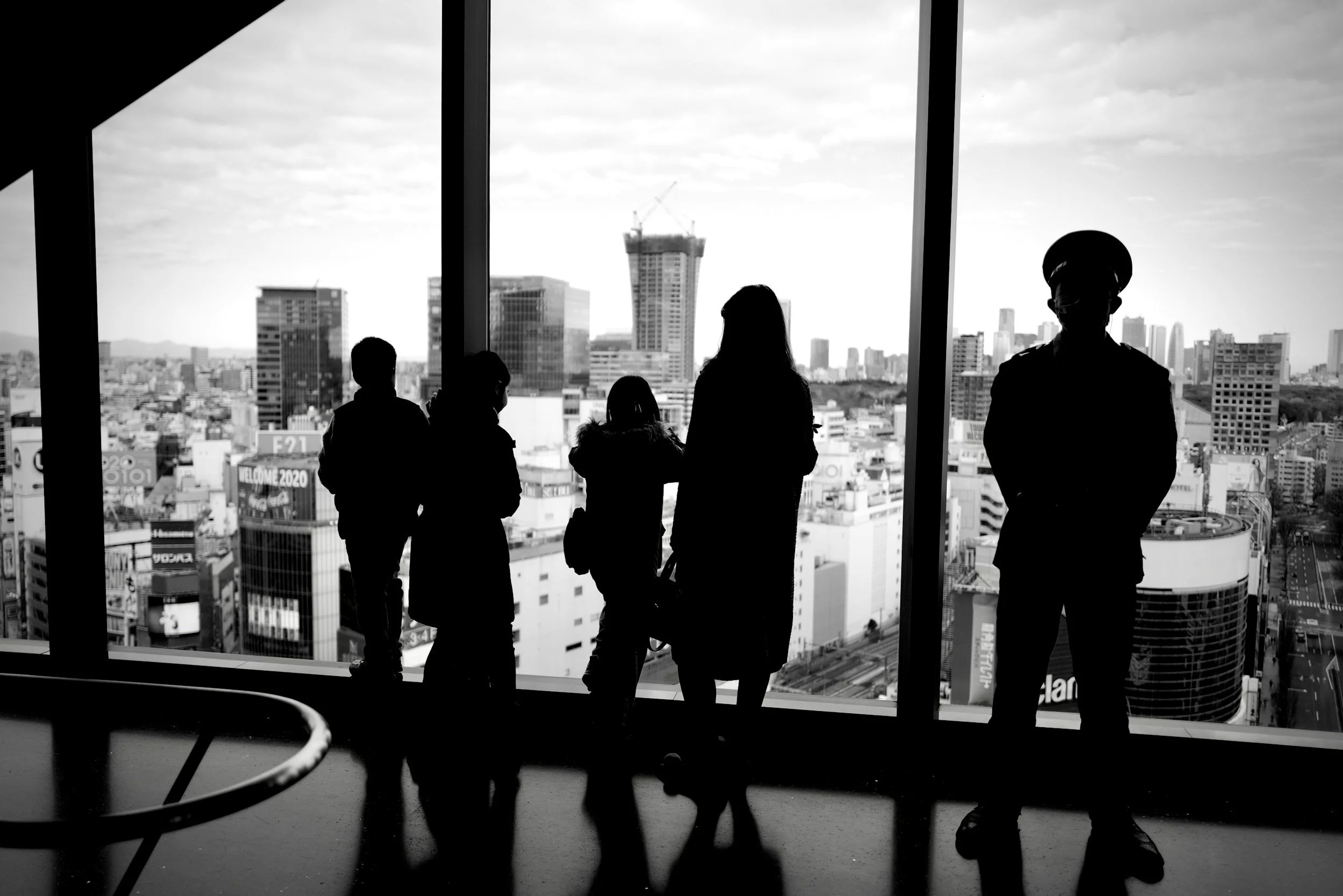 Silhouettes of five people looking out a large window at a city skyline with tall buildings, one under construction, in black and white.