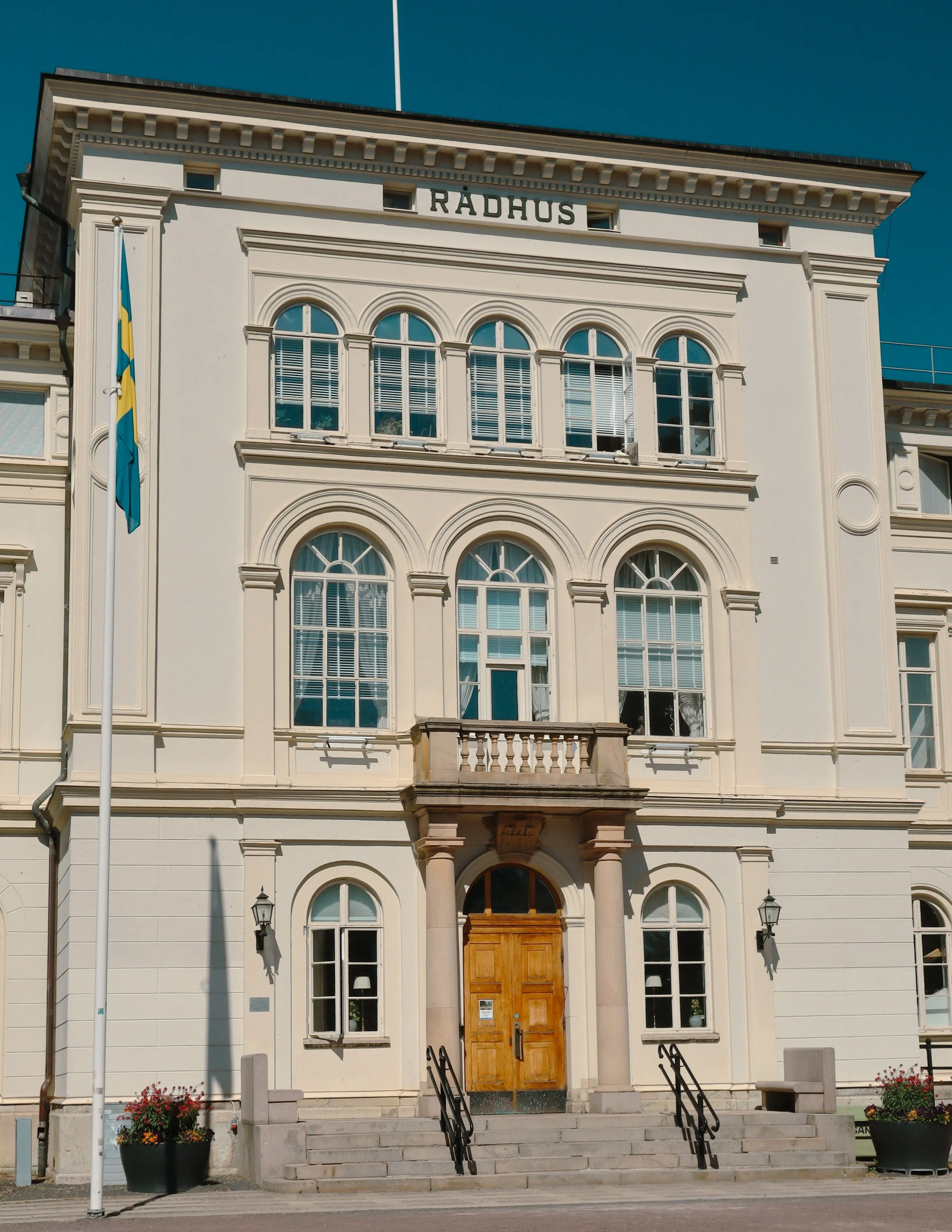 Front view of a government building with a sign that reads 'RÄDHUS', featuring a wooden main door, large arched windows, steps leading up to the entrance, and a flagpole with a flag on the left side.