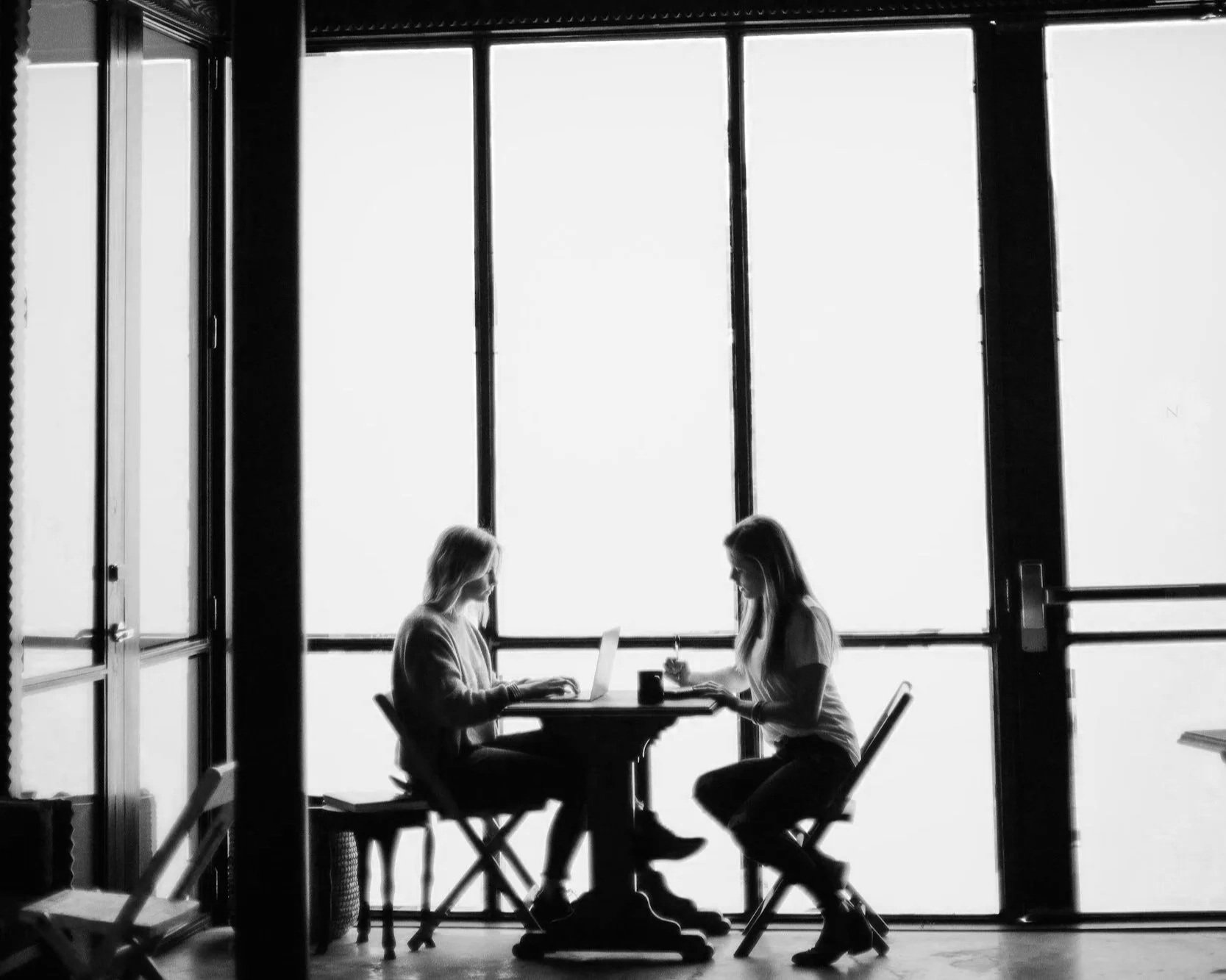Two women sit at a table in front of a large window, working on laptops in a bright, modern space with natural light.