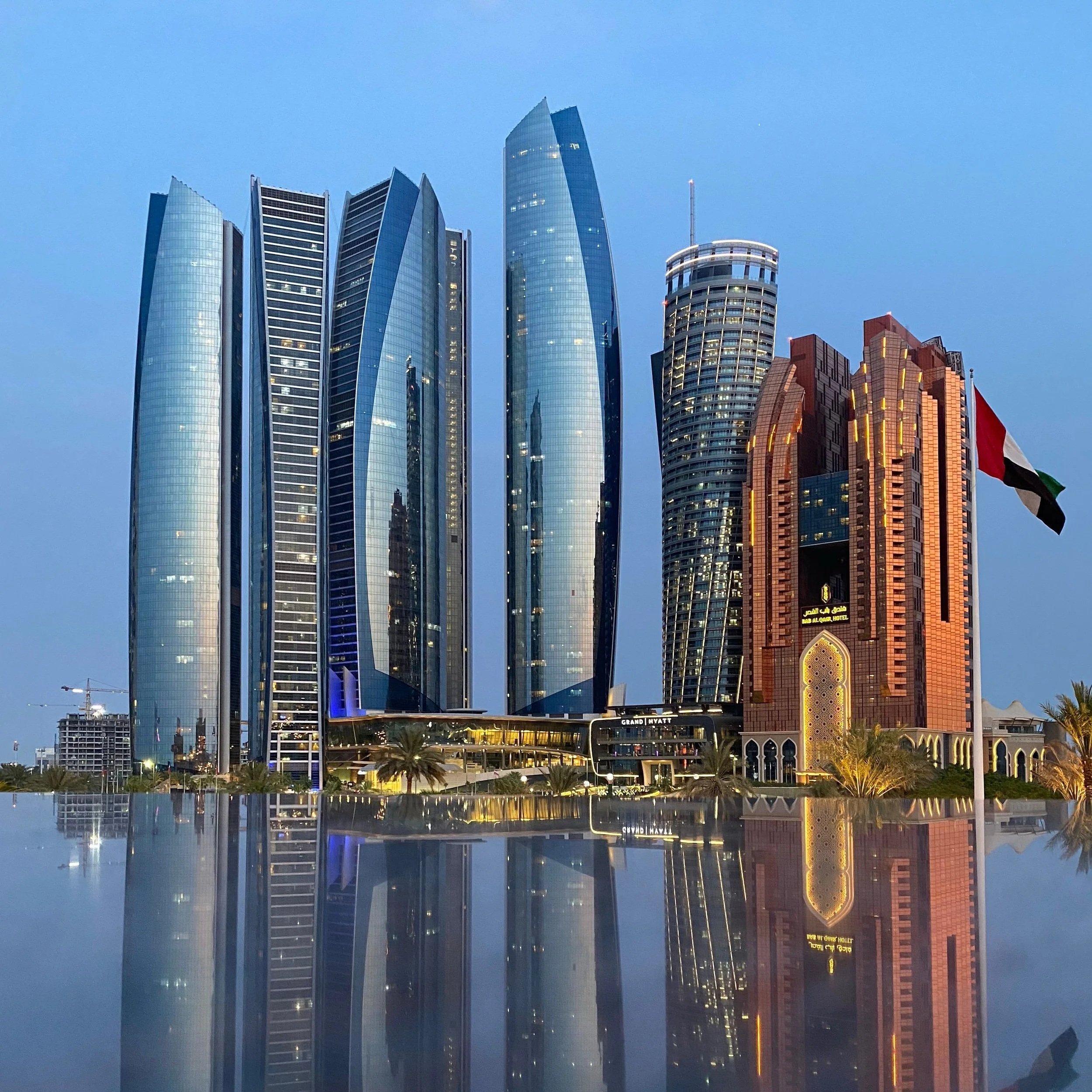 Tall modern skyscrapers with glass facades reflecting the sky, buildings illuminated with warm lights, Dubai skyline at dusk, with a pool and palm trees in the foreground.