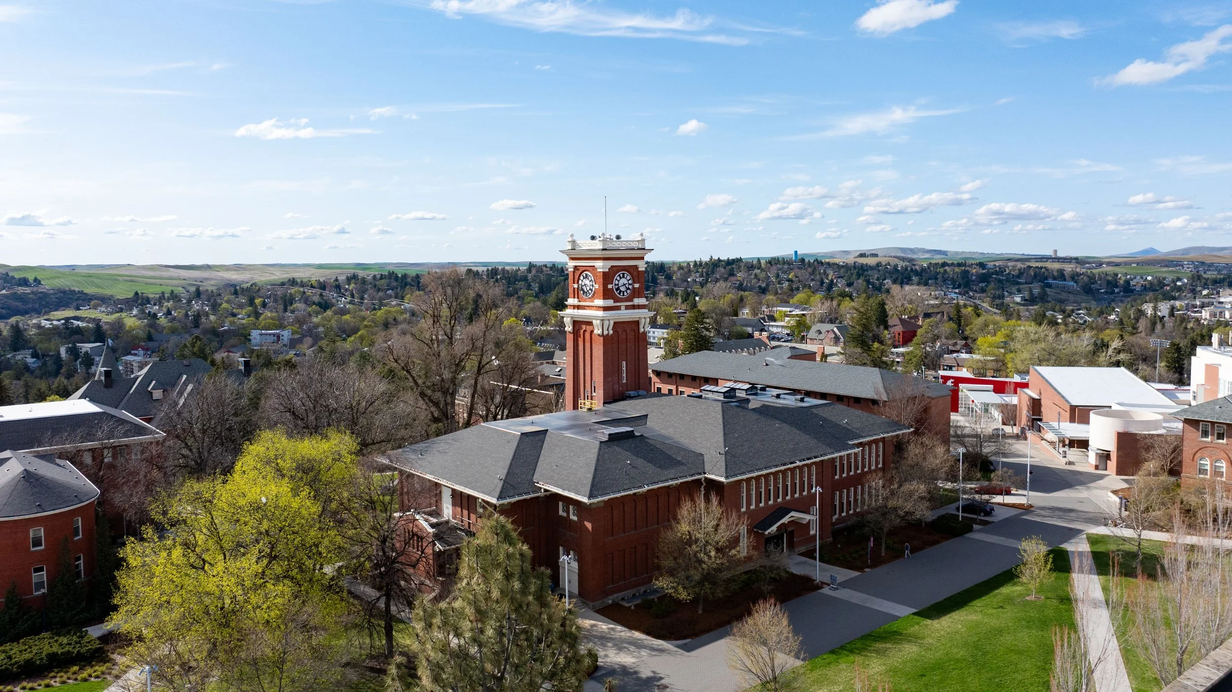 Aerial view of a town with a prominent red brick clock tower, surrounded by trees and buildings, with hills and fields in the background under a partly cloudy sky.