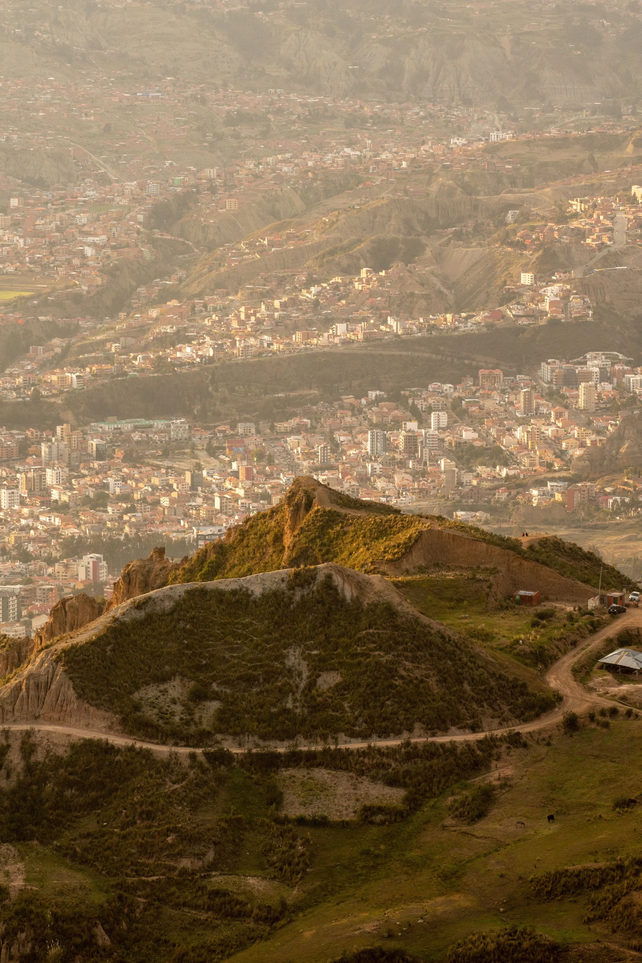 A cityscape with densely packed buildings in a valley between mountains, with a winding dirt road in the foreground and a hill at the bottom of the image.