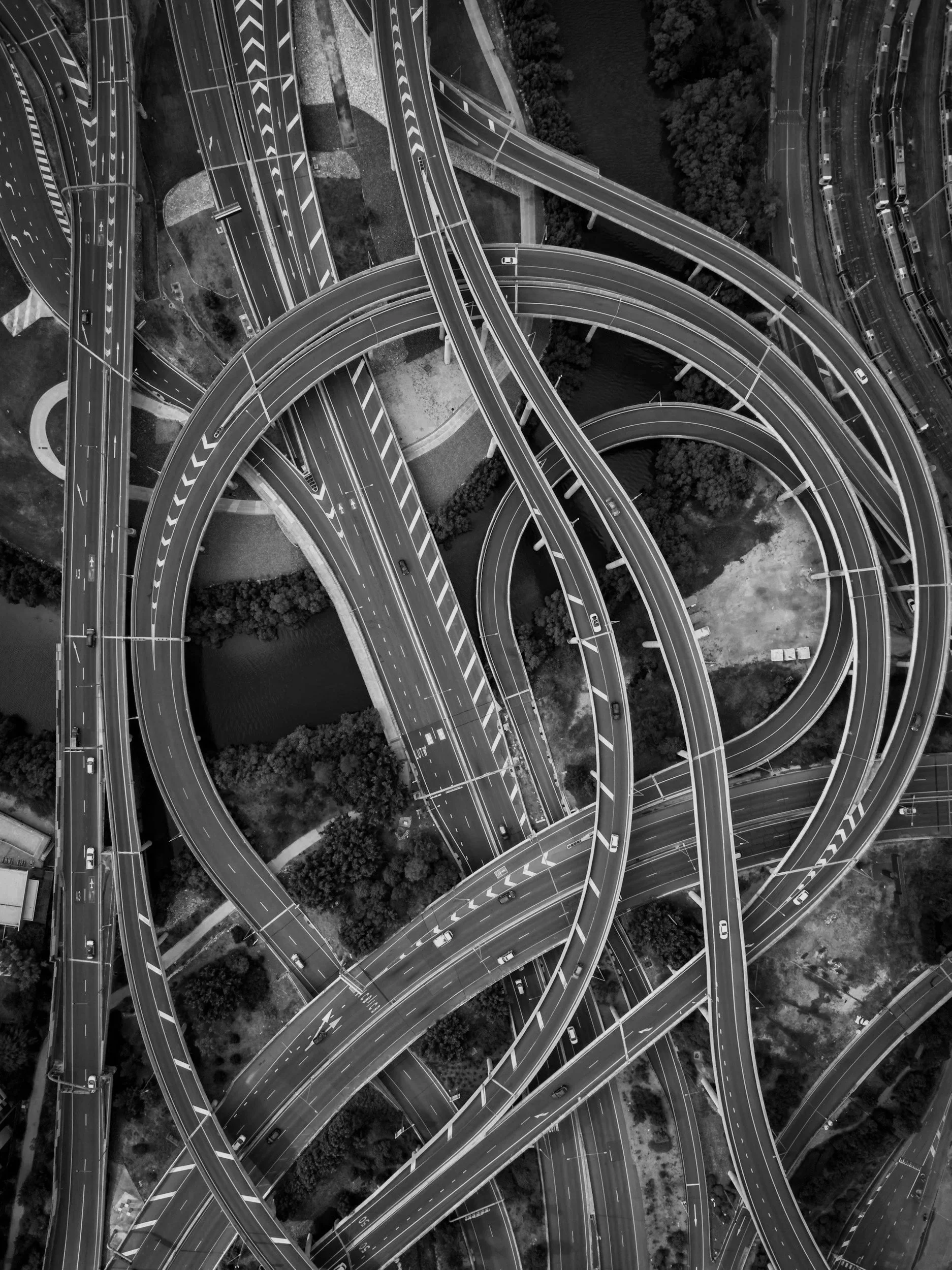 Aerial view of a multi-level highway interchange with multiple looping and curved ramps, vehicles, and road signs, surrounded by greenery and water.