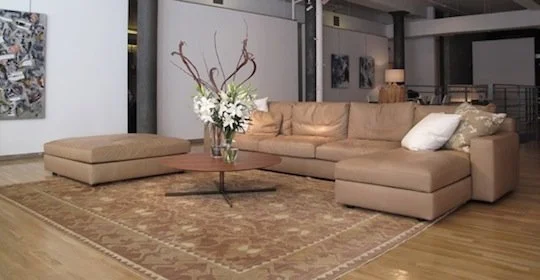 Living room with beige sectional sofa, glass coffee table with floral arrangement, and a patterned area rug.