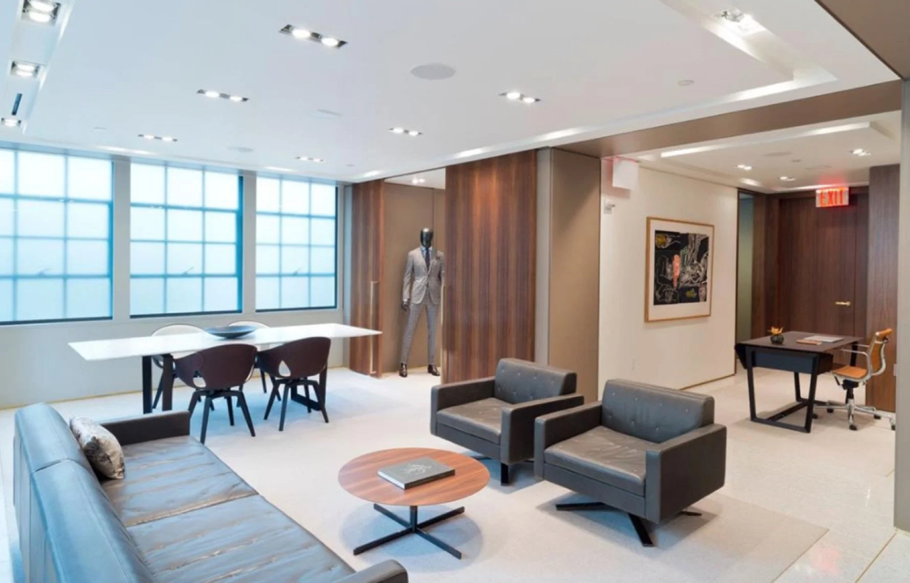 Modern lounge area with sofa, armchairs, coffee table, dining table with chairs, and a wooden mannequin dressed in a suit in the corner, next to large windows and contemporary artwork.
