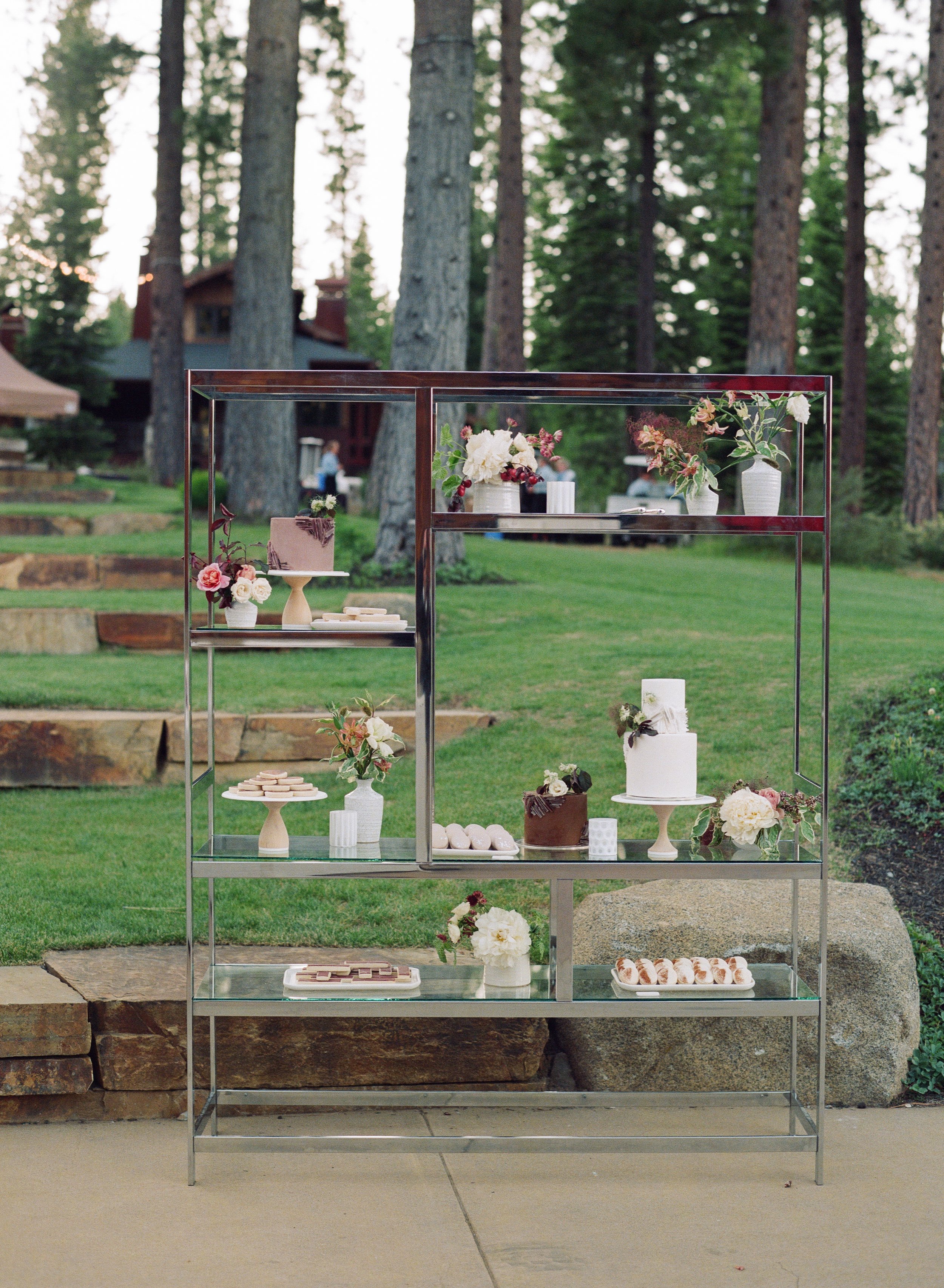 Display stand with floral decorations and desserts set outdoors in a green, wooded area.