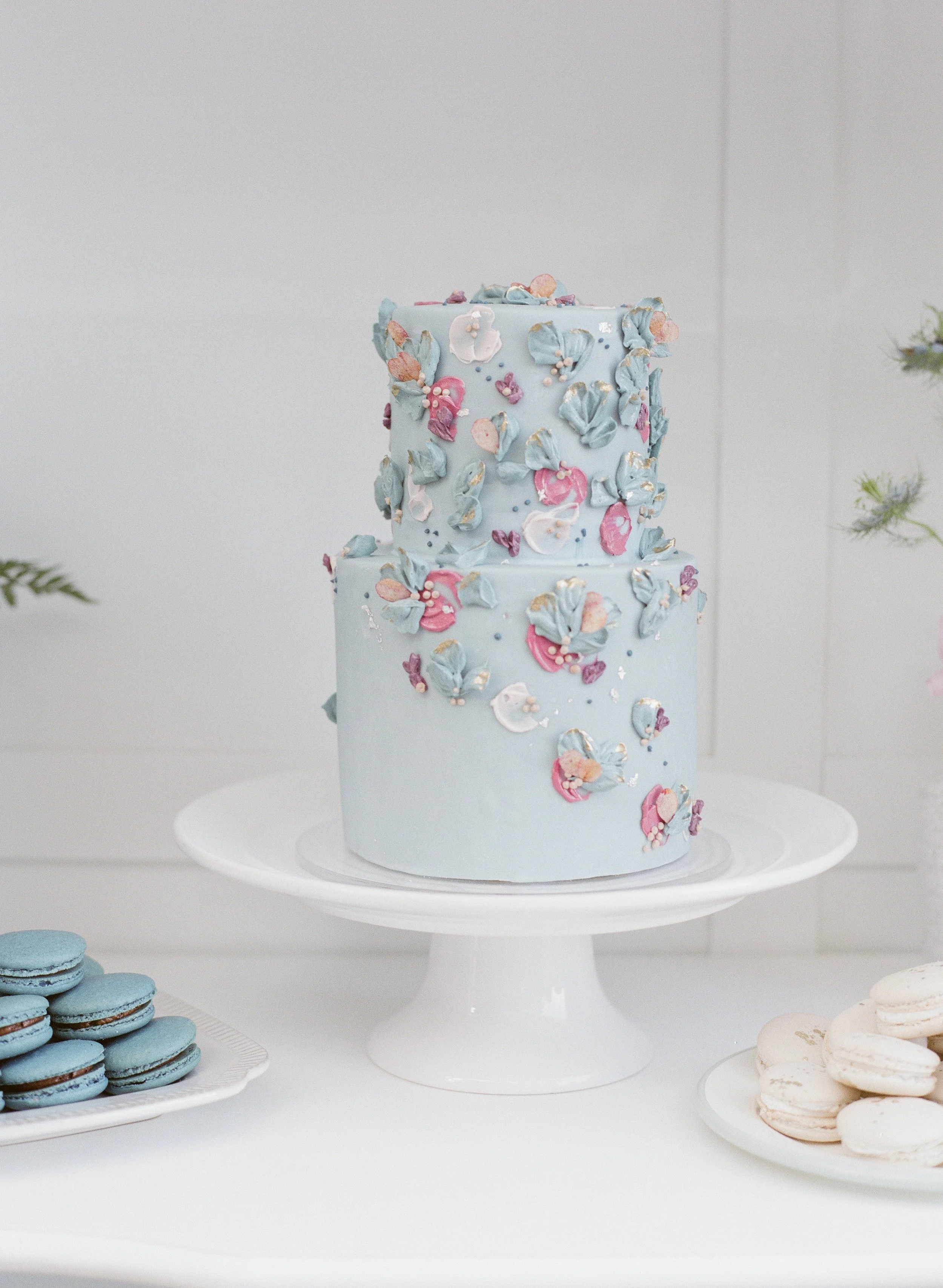Two tiered blue cake decorated with pastel-colored flower and leaf piped frosting and metallic accents, displayed on a white cake stand. Surrounding desserts include blue macarons and white cookies.