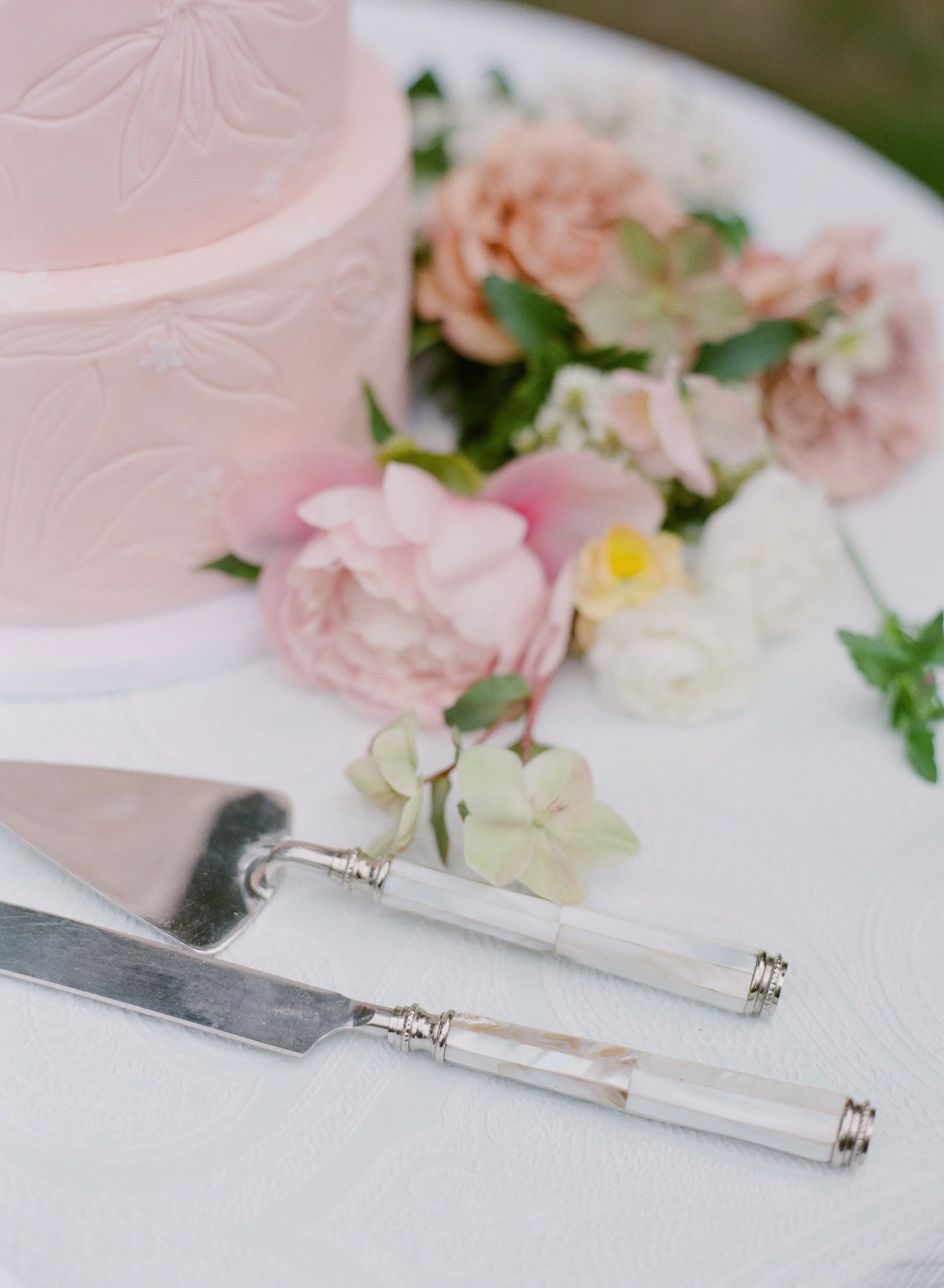 A pink tiered wedding cake with embossed floral designs, decorated with pink and peach flowers and green leaves, on a white surface. Two vintage silver-handled knives are placed in front of the cake.