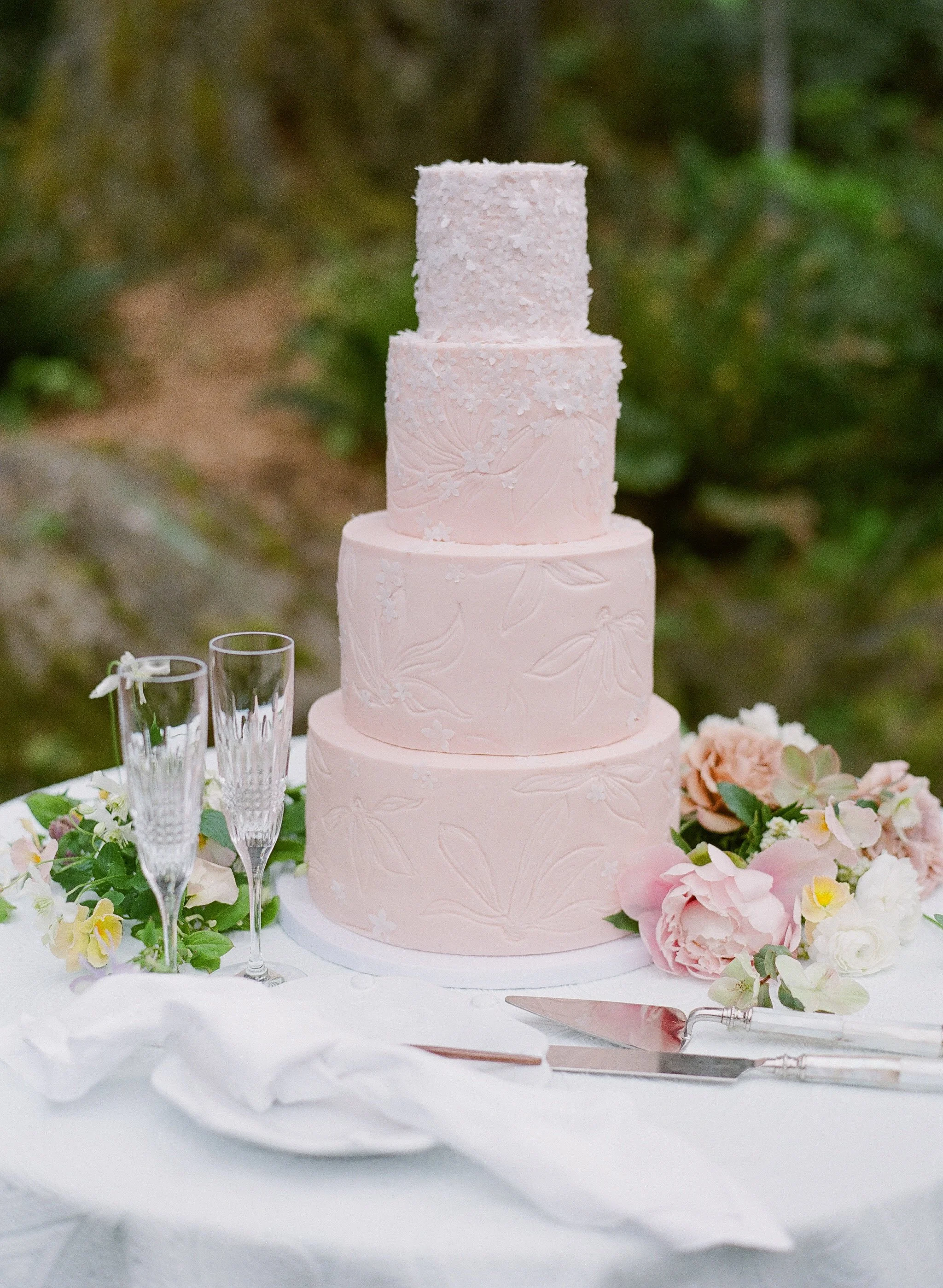 A pink, multi-tiered wedding cake decorated with floral patterns and small white flowers, placed on a table with flowers and glassware.