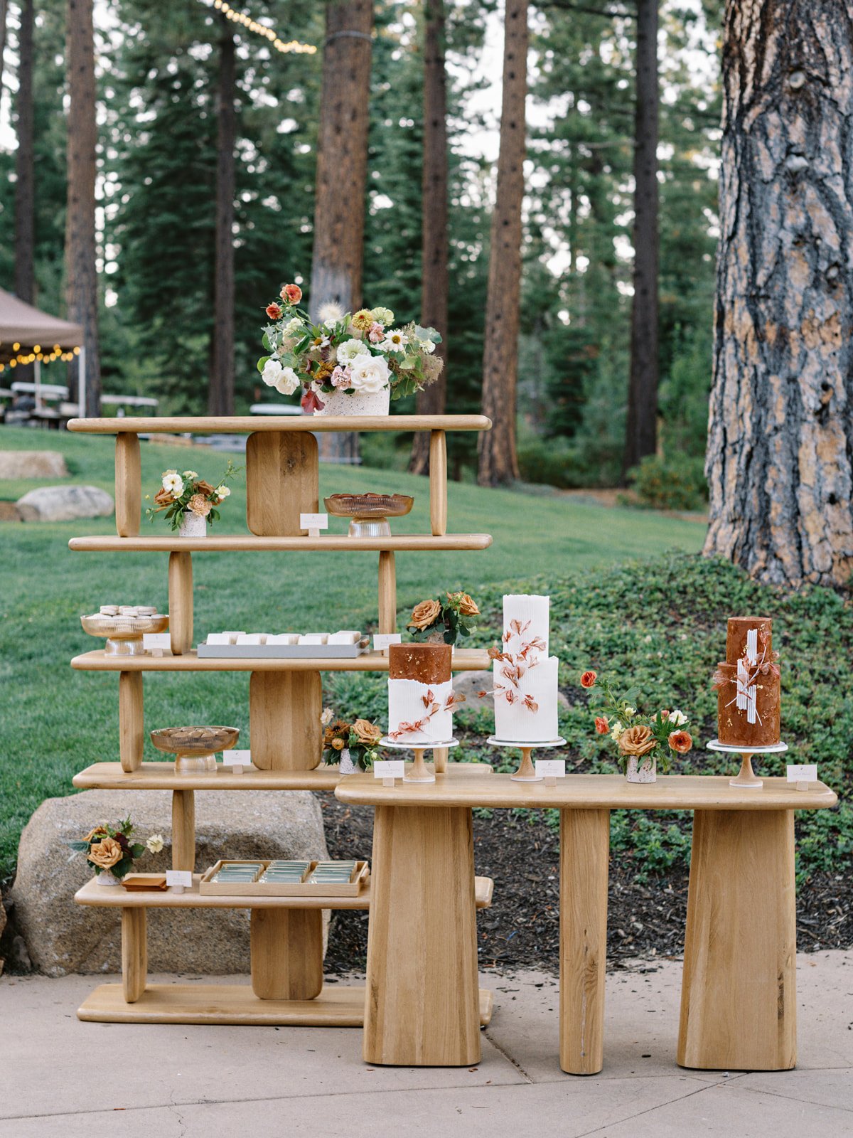 Display of floral arrangements, candles, and baked goods on wooden shelves in an outdoor setting surrounded by trees.