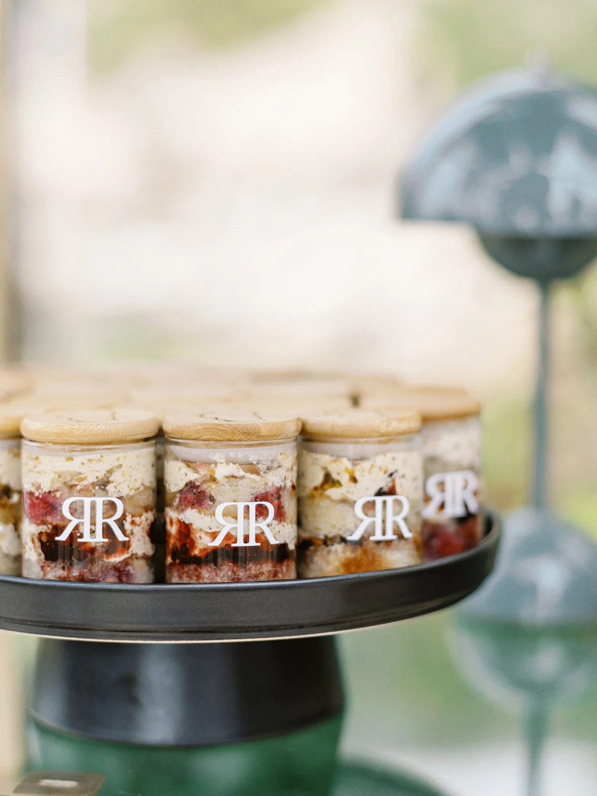A round black platter with several small glass jars of layered dessert with a creamy topping, strawberries, and other fruit, labeled with white logos featuring two capital R's. In the background, there is a blurred outdoor scene with a blue grill.