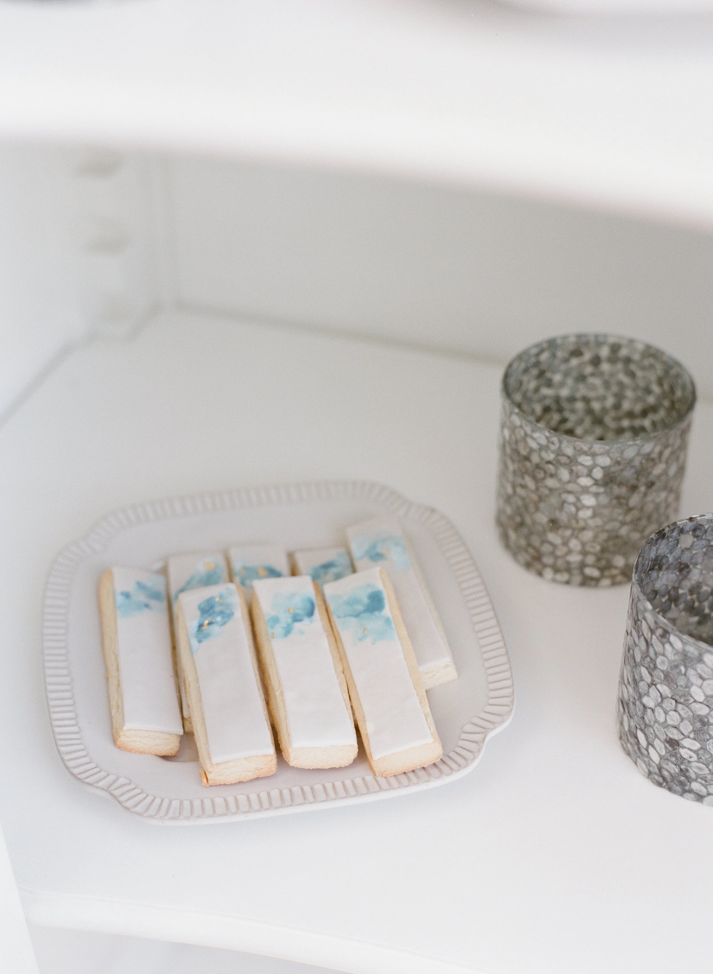A white tray with cookies decorated with blue watercolor patterns and small toppings, two mosaic-patterned vases, and a white candlestick holder with small candles.